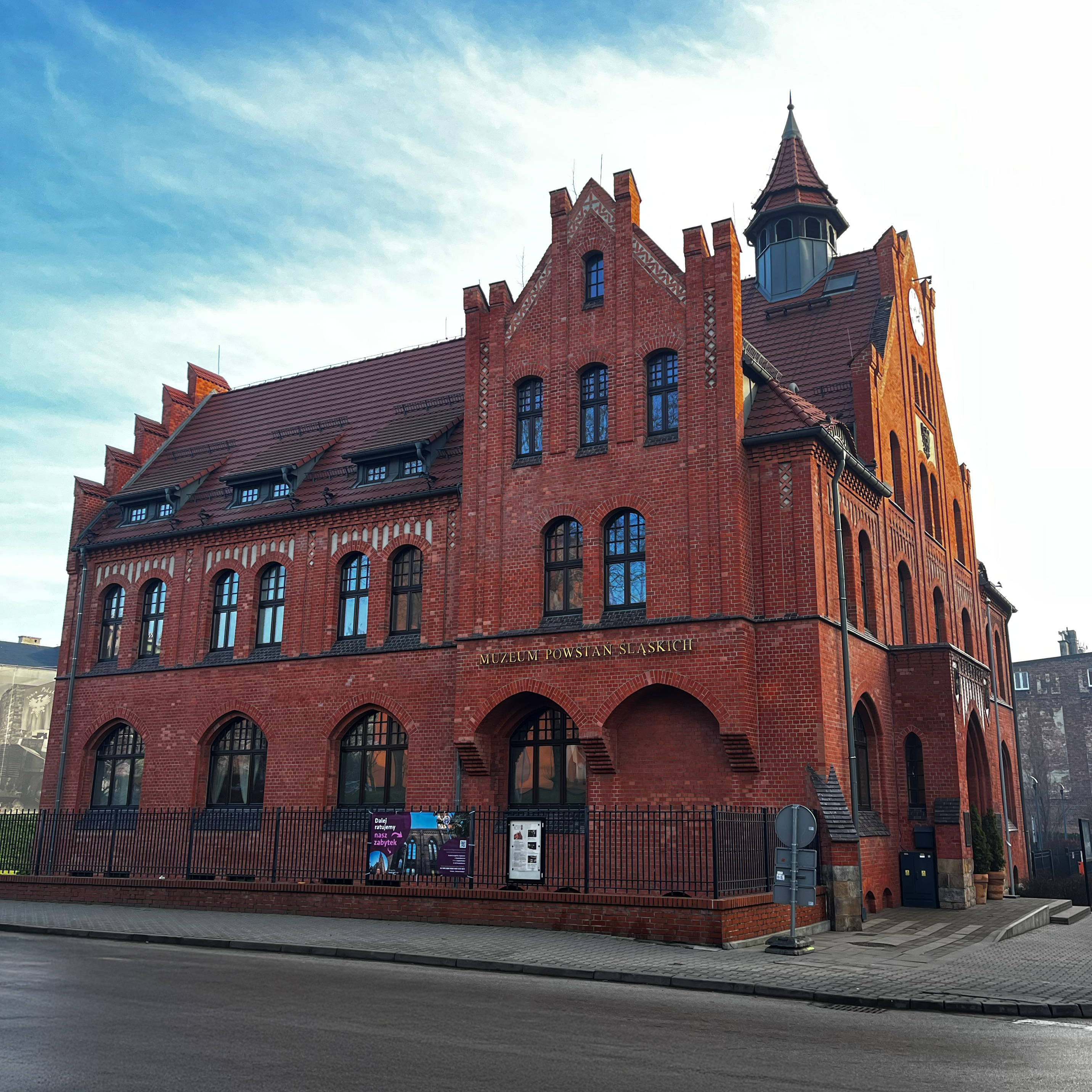 "Ceglany budynek Muzeum Powstań Śląskich w Świętochłowicach, o neogotyckiej architekturze z wieżyczką i ozdobnymi szczytami. Na fasadzie widoczny złoty napis z nazwą muzeum. Budynek otoczony czarnym ogrodzeniem, w tle niebo z lekkim zachmurzeniem." },