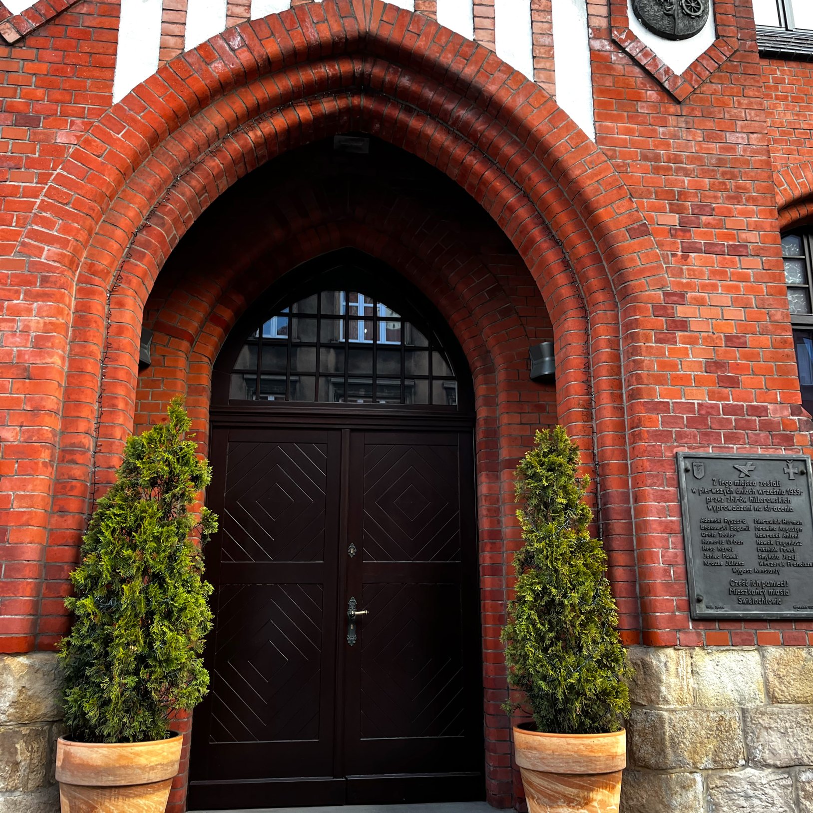 "Zbliżenie na wejście do Muzeum Powstań Śląskich z łukowym ceglanym portalem, dużymi drewnianymi drzwiami oraz dwoma doniczkami z iglakami po bokach. Obok wejścia umieszczona jest tablica pamiątkowa."