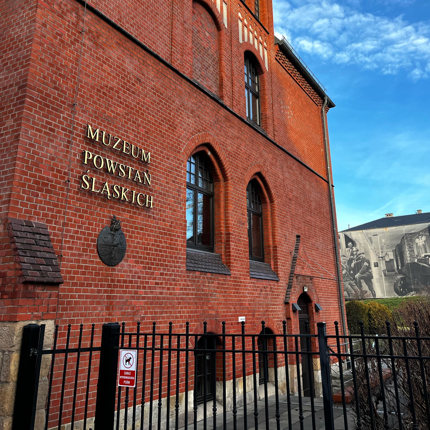 "Fragment ściany Muzeum Powstań Śląskich z widocznym napisem w złotych literach oraz herbem. Ceglany mur, ostrołukowe okna i fragment metalowego ogrodzenia na pierwszym planie. W tle mural z archiwalnym zdjęciem powstańców na ścianie sąsiedniego budynku."