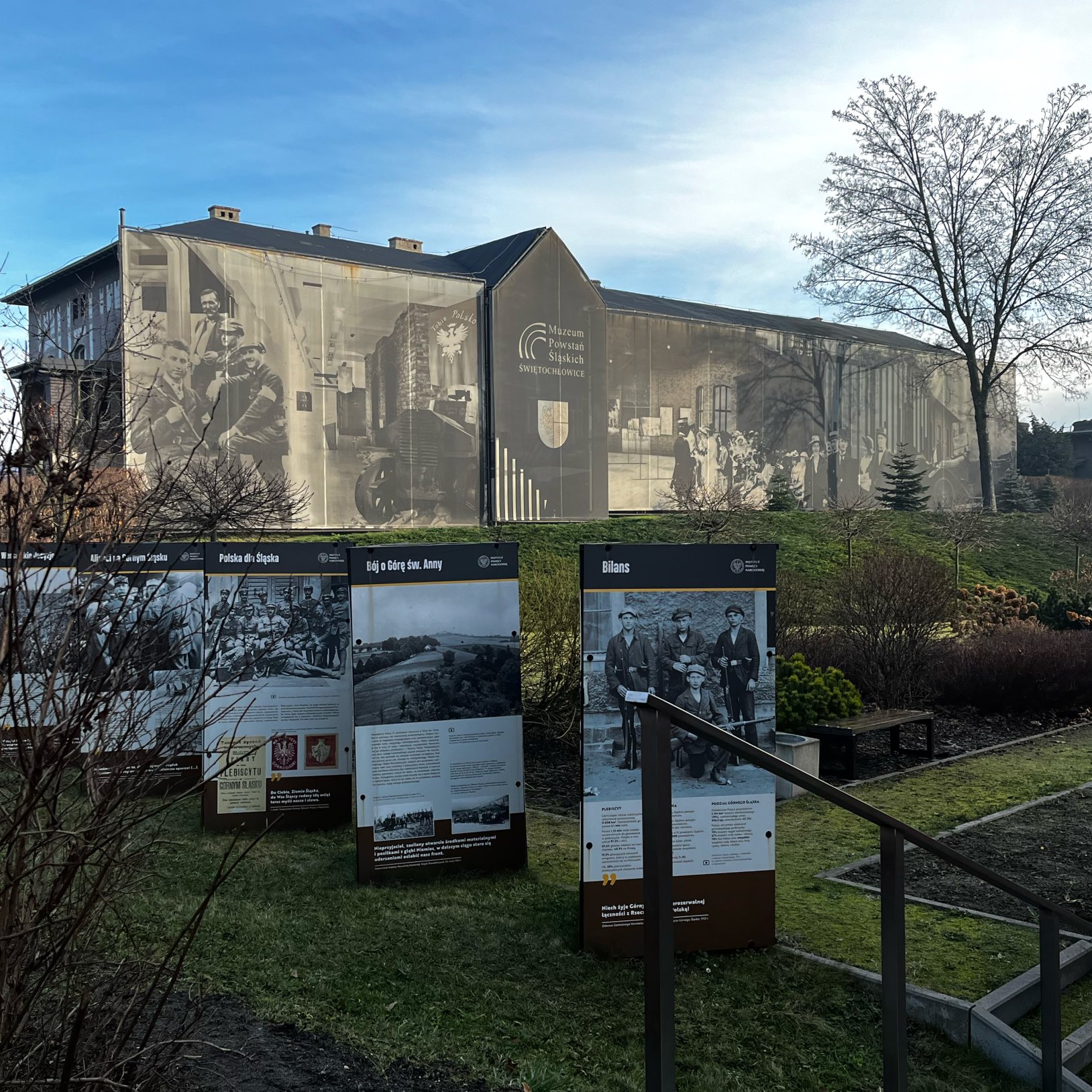 "Ekspozycja plenerowa przy Muzeum Powstań Śląskich przedstawiająca plansze informacyjne z archiwalnymi zdjęciami i opisami. W tle mural ze scenami historycznymi oraz logotypem muzeum na ścianie sąsiedniego budynku. Otoczenie zieleni i bezlistnych krzewów.