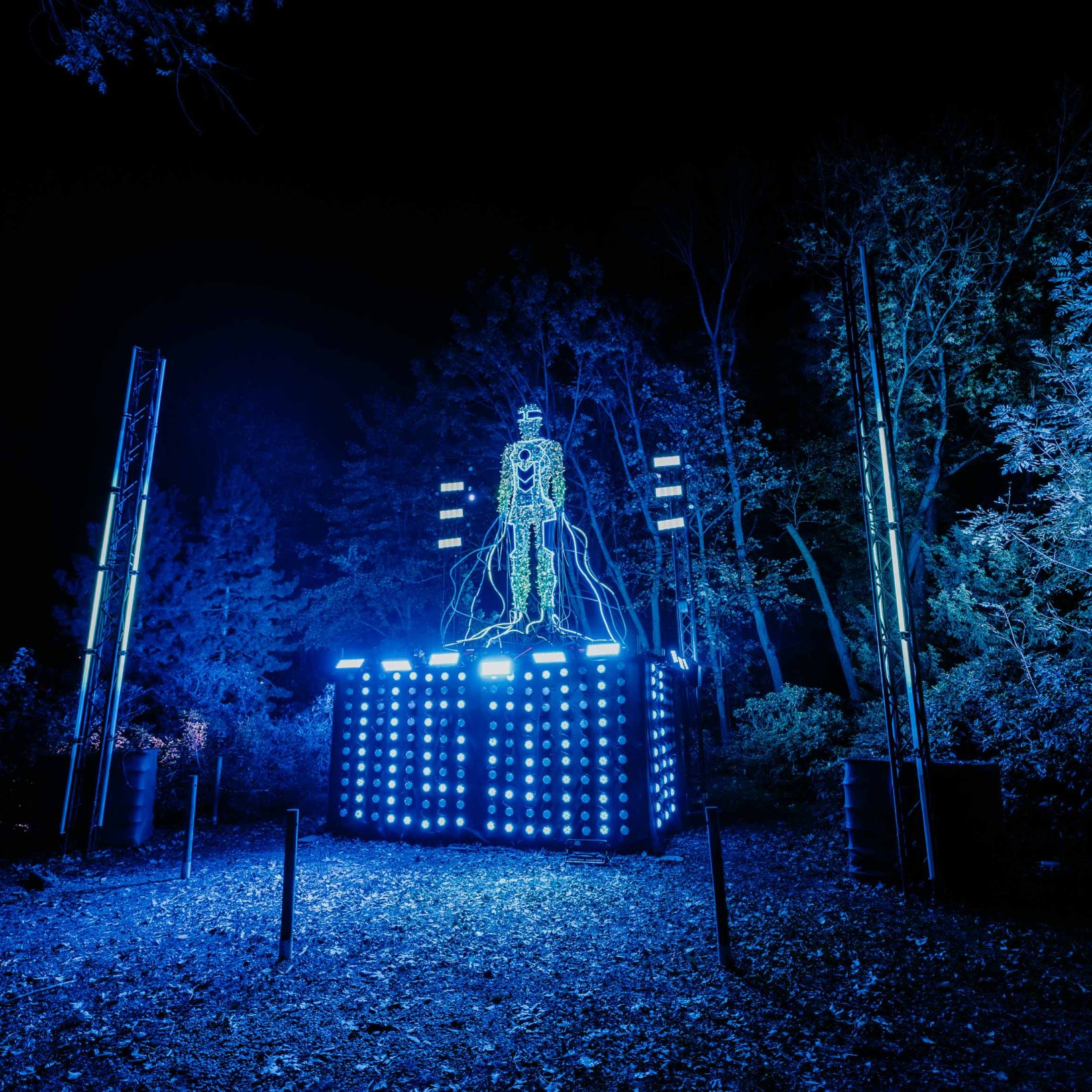 "Instalacja świetlna przedstawiająca postać ludzką zbudowaną z kabli i świateł, ustawiona na dużym podeście podświetlonym intensywnym niebieskim światłem. Wokół znajduje się las, również oświetlony na niebiesko. Zdjęcie wykonane nocą."   },