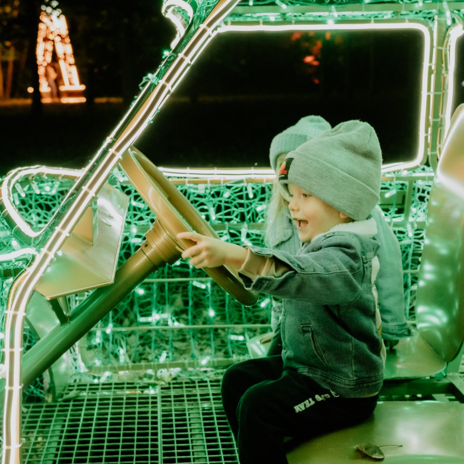 "Dwójka dzieci w czapkach i kurtkach siedzi w ozdobnej instalacji w kształcie samochodu, podświetlonej jasnozielonymi światłami LED. Jedno z dzieci trzyma kierownicę i z entuzjazmem udaje jazdę. W tle widoczne są kolorowe dekoracje świetlne w parku."