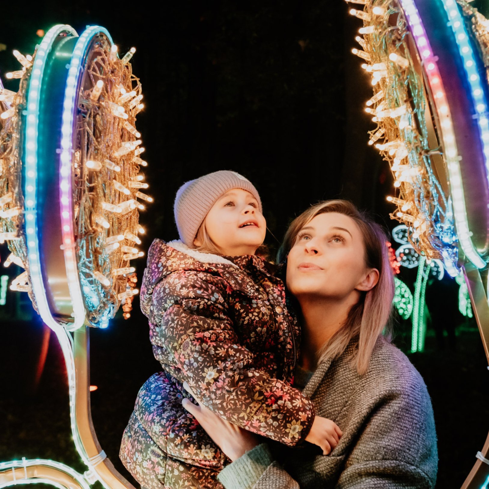 "Kobieta trzyma na rękach małą dziewczynkę w kurtce w kwiaty i szarej czapce. Oboje patrzą w górę na kolorową iluminację w kształcie dużych kwiatów z mnóstwem światełek LED. Scena ma miejsce nocą w parku z licznymi świetlnymi dekoracjami w tle."