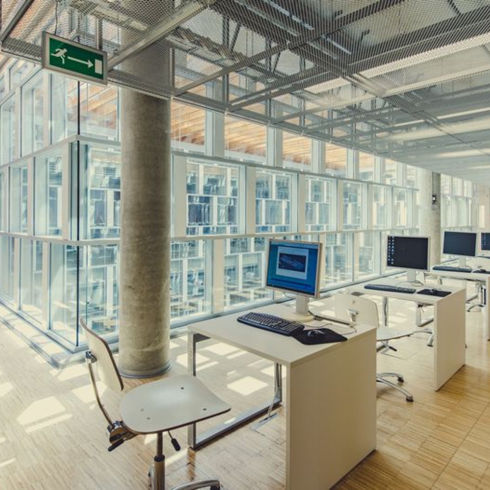 Wnętrze biblioteki z rzędami biurek przy przeszklonej ścianie. Na biurkach znajdują się komputery, a przy nich białe krzesła. W tle widoczna nowoczesna, przeszklona konstrukcja budynku.