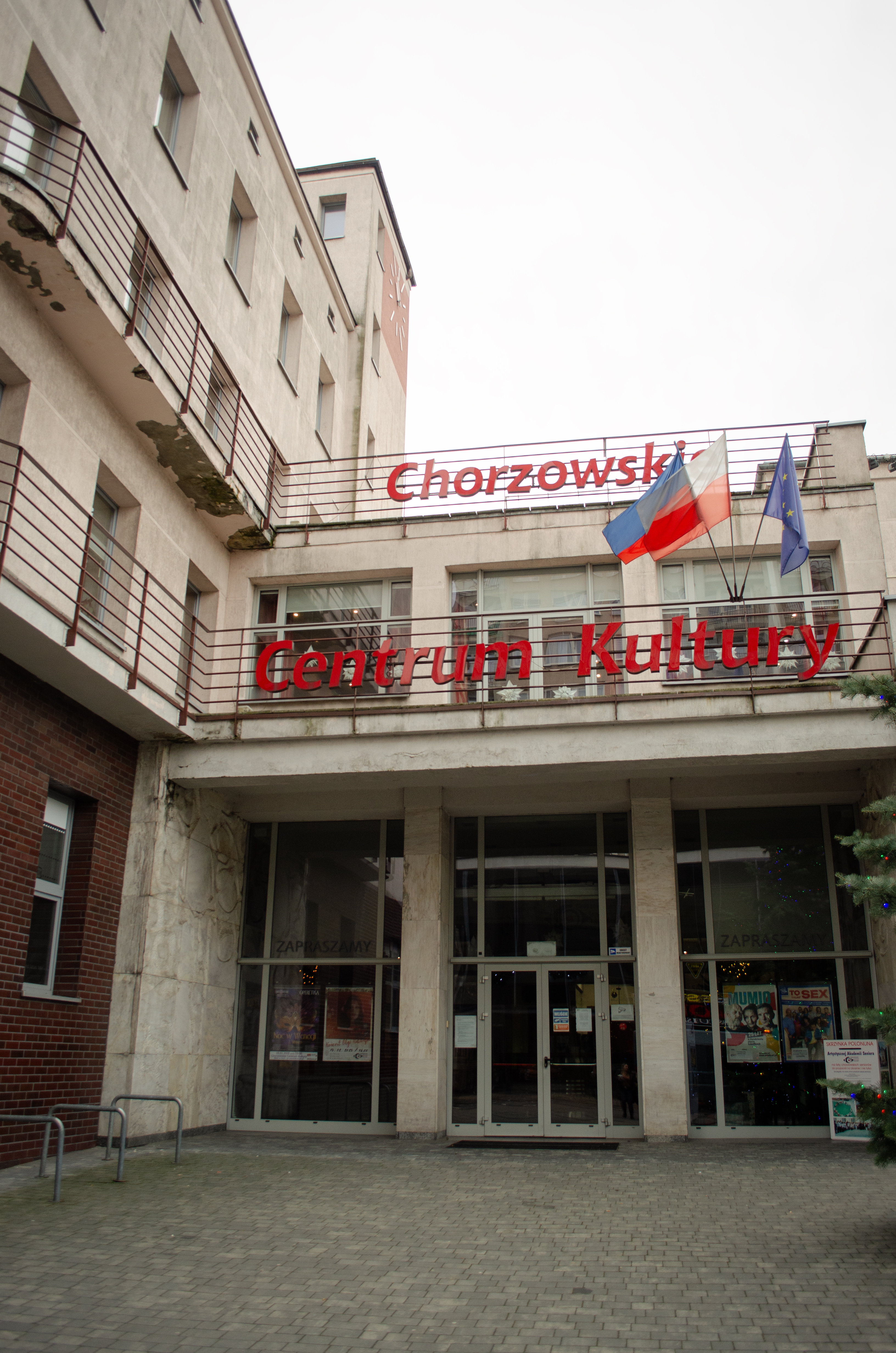 "Wejście do Chorzowskiego Centrum Kultury, widoczne z dołu pod lekkim kątem. Na fasadzie budynku znajduje się czerwony napis „Chorzowskie Centrum Kultury” oraz trzy flagi: Polski, województwa śląskiego i Unii Europejskiej. Budynek ma jasną elewację i duże