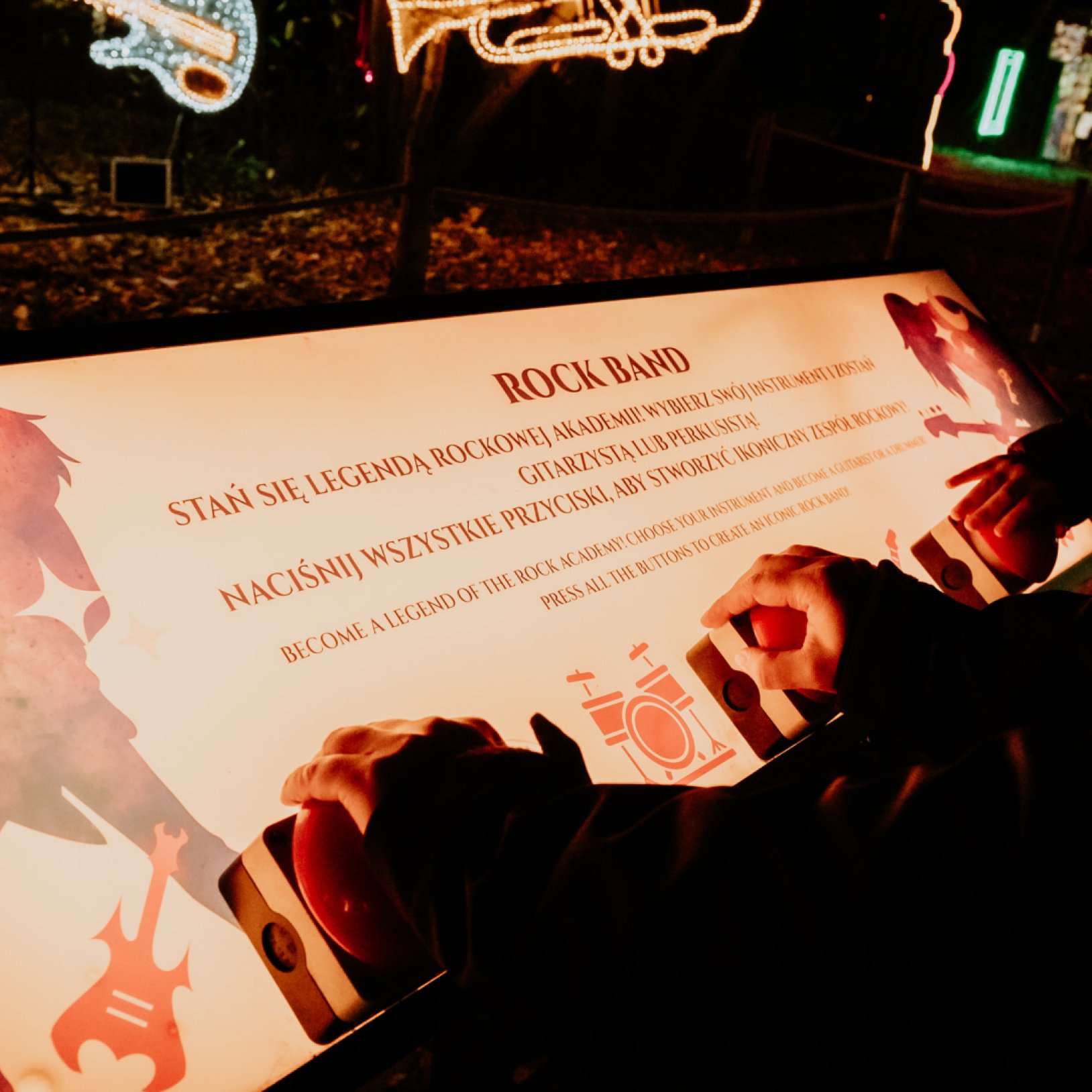"Zbliżenie na tablicę interaktywną z napisem 'Rock Band', z instrukcjami po polsku i angielsku. Dzieci naciskają przyciski na tablicy, a w tle widoczne są iluminacje w kształcie instrumentów muzycznych, takich jak gitara i trąbka."