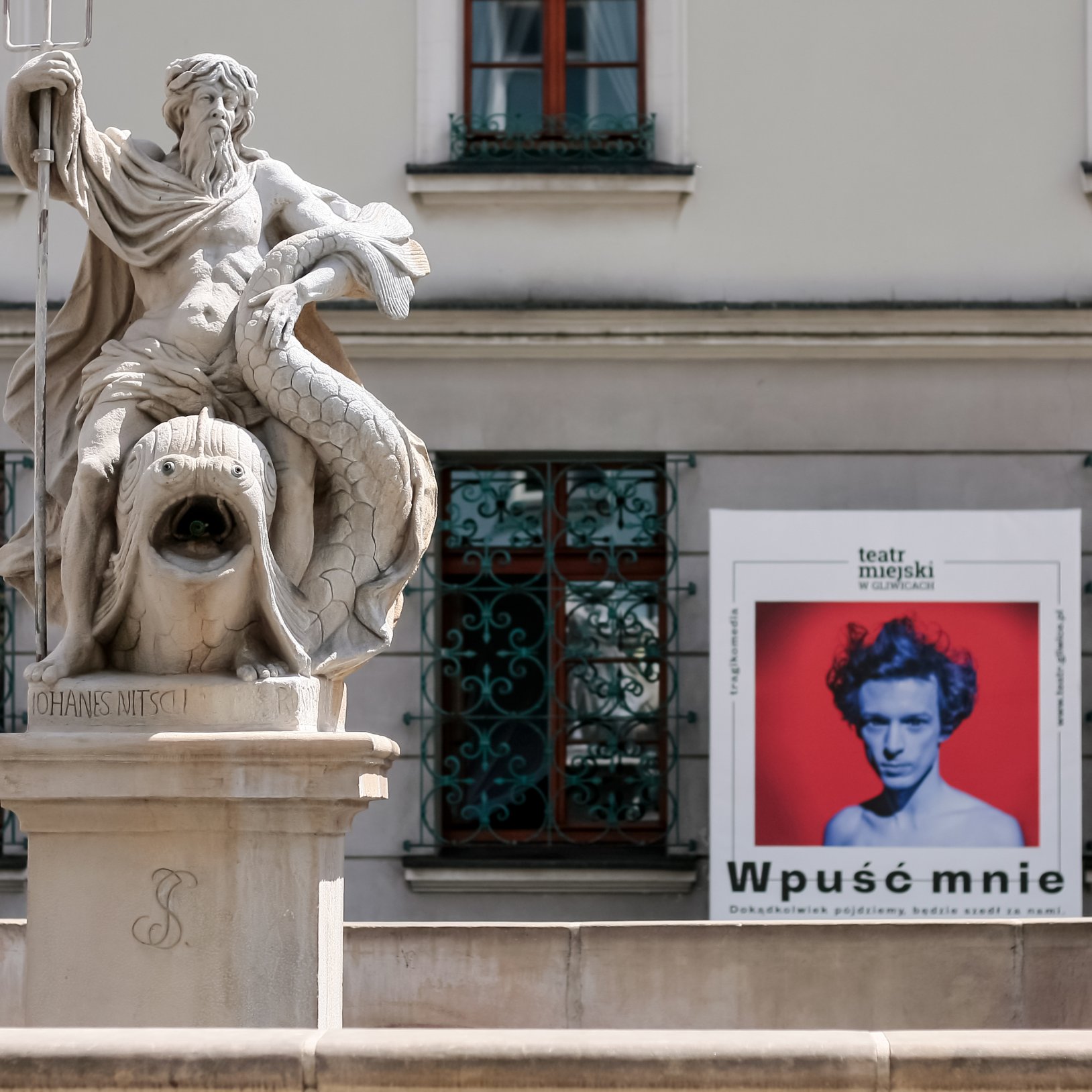 "Rzeźba Neptuna z trójzębem, siedzącego na rybie, umieszczona na fontannie. W tle znajduje się budynek z kratowanymi oknami i plakatem spektaklu 'Wpuść mnie' Teatru Miejskiego w Gliwicach, przedstawiającym osobę z czerwonym tłem."