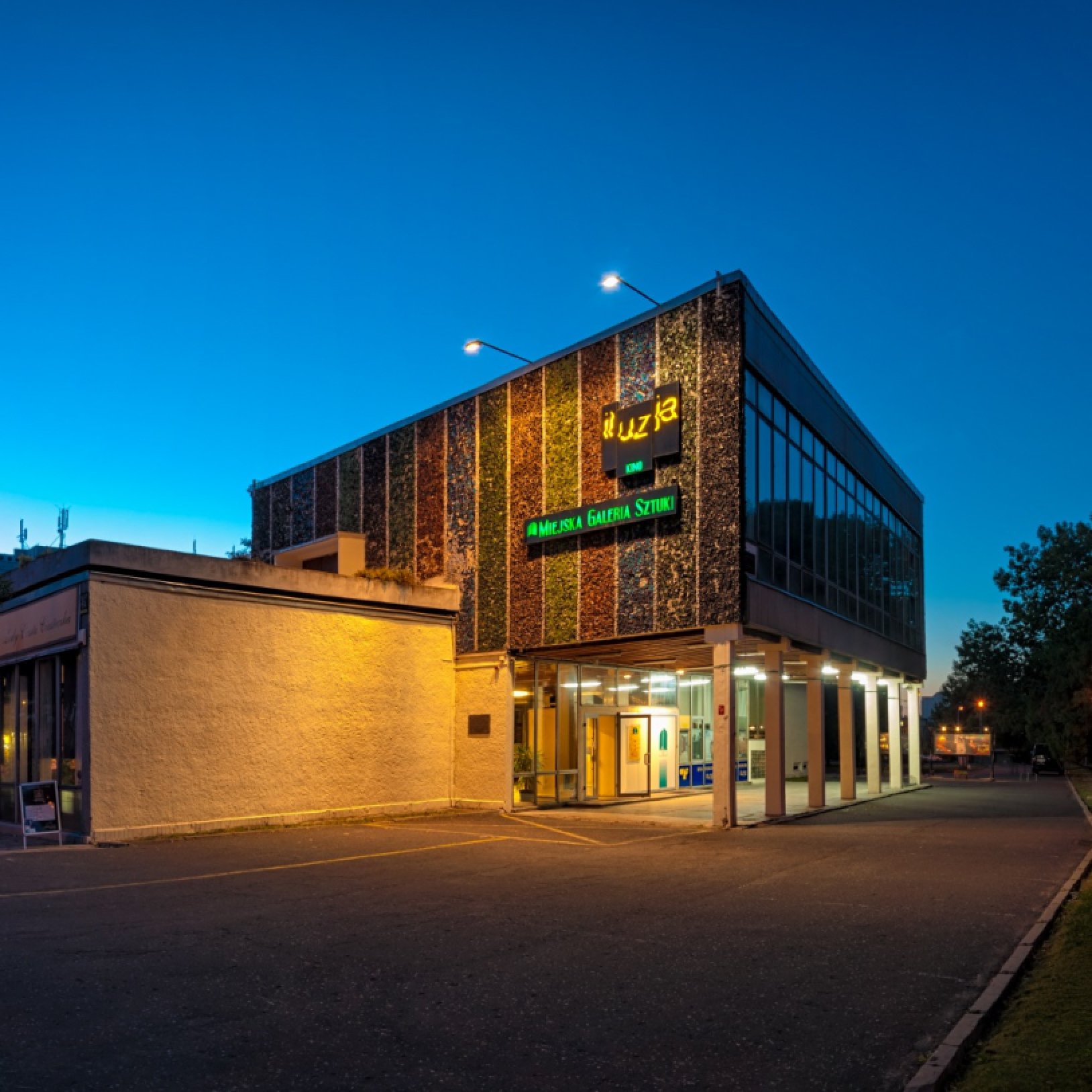 Budynek kina o elewacji z pionowych pasów mozaiki, oświetlony po zmroku; nad wejściem świecą napisy „Iluzja” i „Miejska Galeria Sztuki”, a z boku widać przeszkloną część i podcienie na słupach.