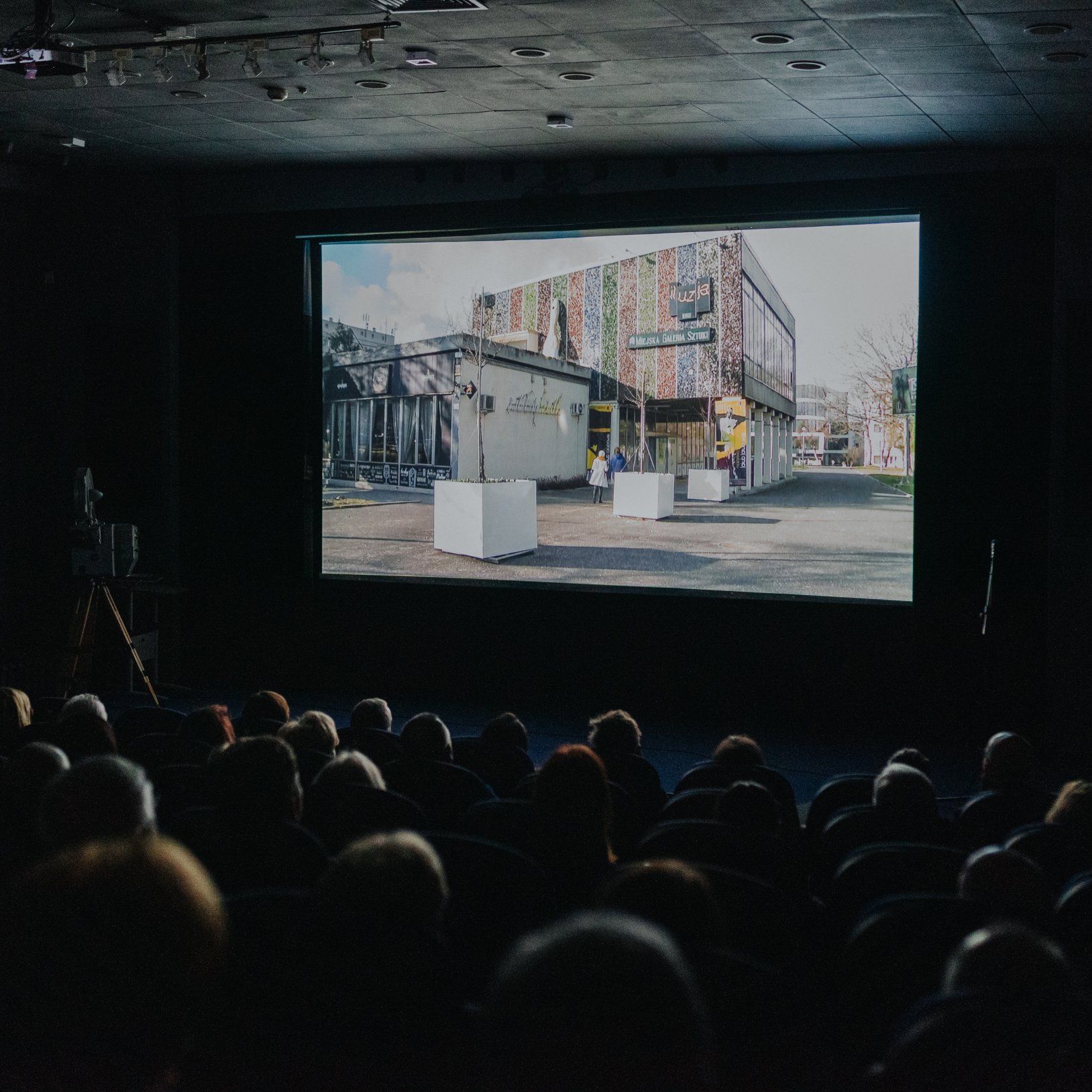 Ciemna sala kinowa z widownią oglądającą projekcję; na dużym ekranie wyświetlony jest budynek z napisem „Iluzja” i „Miejska Galeria Sztuki”, a w tle widać sufit z oświetleniem i sprzętem.
