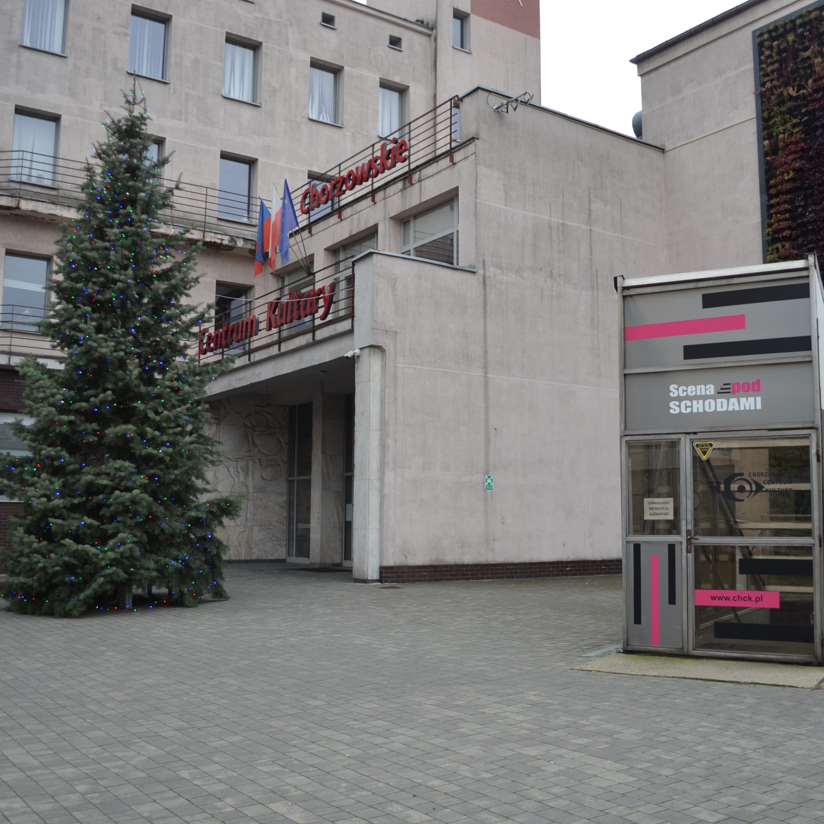 "Plac przed wejściem do Chorzowskiego Centrum Kultury w ciągu dnia. Po lewej stronie znajduje się udekorowana, oświetlona choinka. Nad wejściem widoczny czerwony napis „Chorzowskie Centrum Kultury” oraz flagi. Po prawej stronie zdjęcia widoczna jest przes