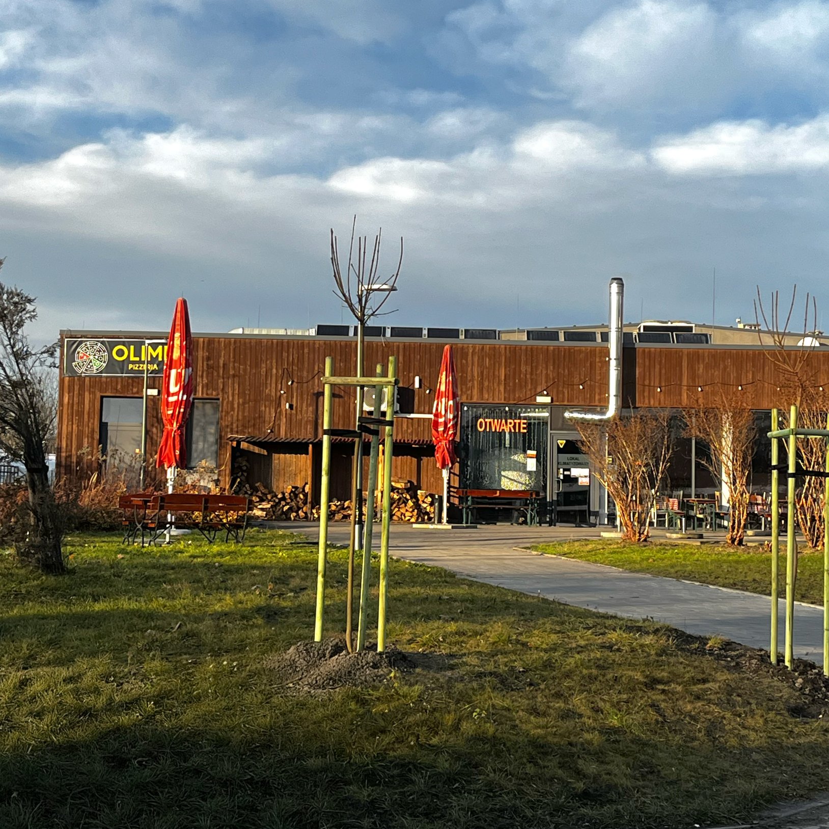 "Budynek pizzerii Olimp z drewnianą elewacją, znajdujący się w otoczeniu zieleni. Przed lokalem stoją czerwone parasole i drewniane ławki, a przy ścianie widać stosy drewna opałowego. Nad wejściem widoczny jest szyld z nazwą lokalu i napisem 'OTWARTE'."