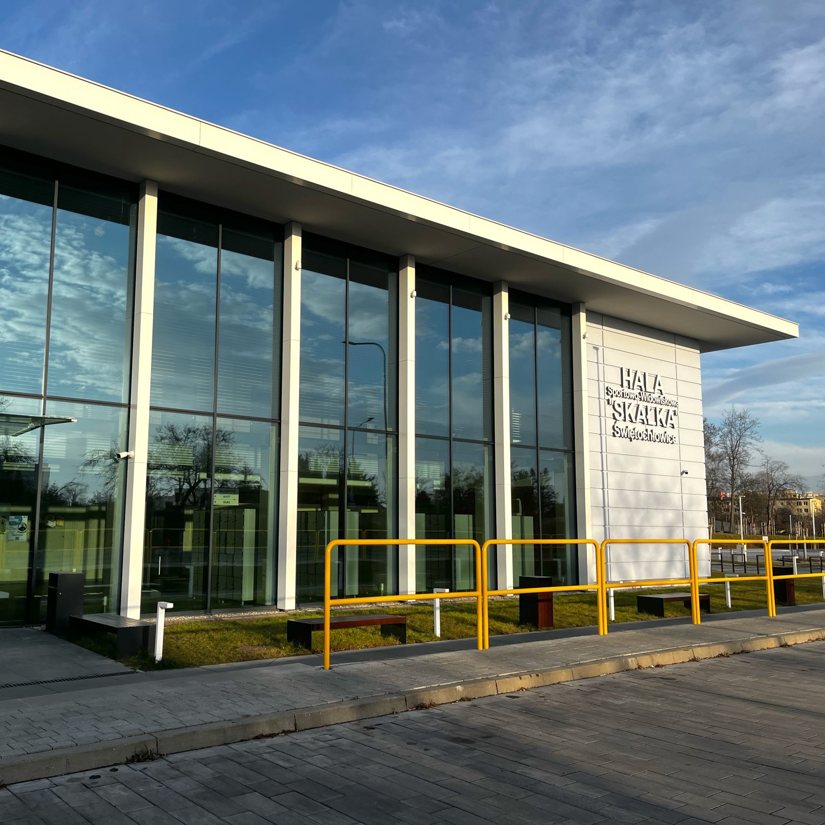 "Fasada Hali Sportowej Skałka w Świętochłowicach z dużymi przeszklonymi oknami i białą elewacją. Na ścianie widnieje napis z nazwą obiektu. Przed budynkiem znajdują się żółte barierki ochronne, ławki i chodnik. W tle widoczne są drzewa i fragmenty miejski