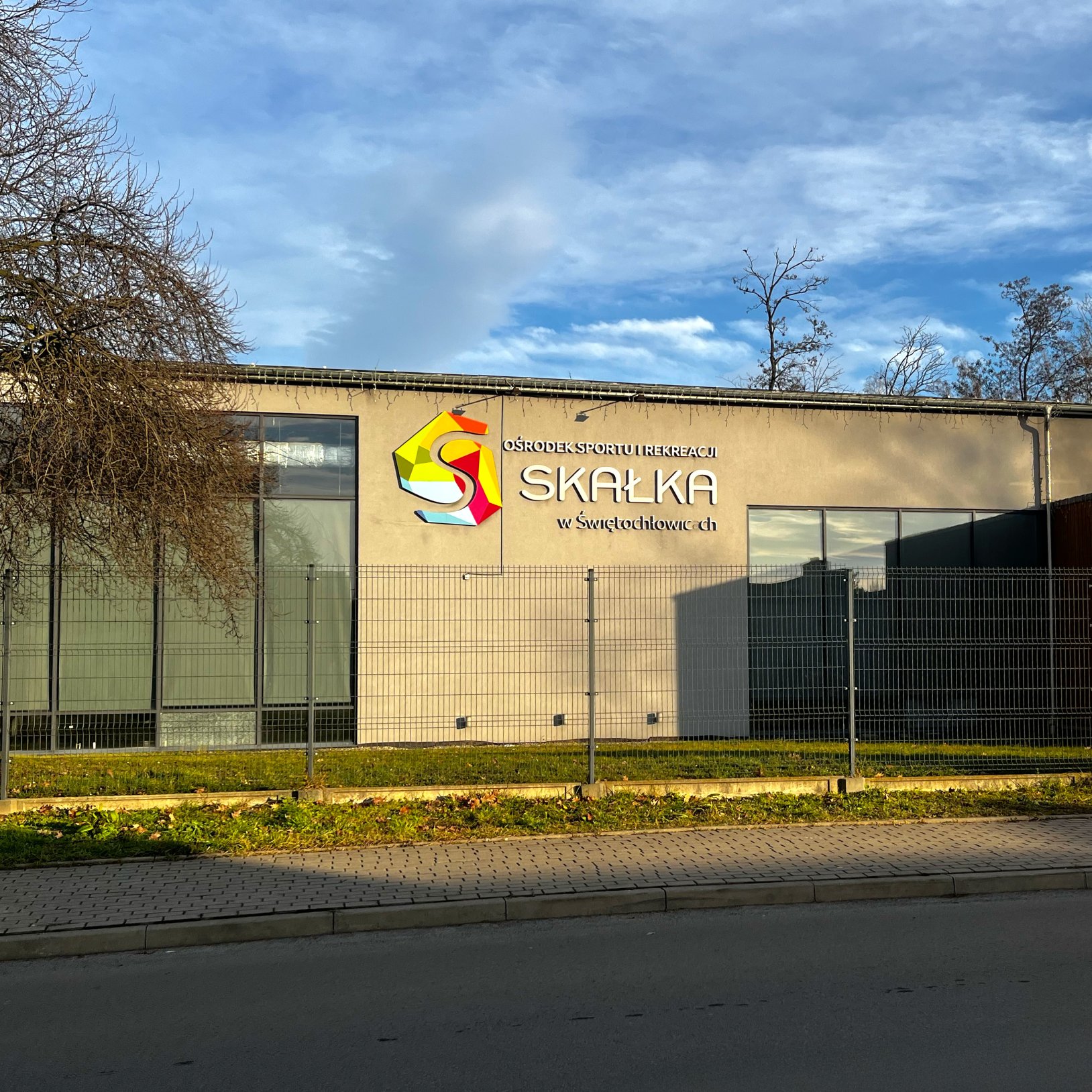 "Widok budynku Ośrodka Sportu i Rekreacji Skałka w Świętochłowicach. Na elewacji znajduje się kolorowe logo i biały napis z nazwą ośrodka. Przed budynkiem widać metalowe ogrodzenie oraz chodnik i fragment jezdni. W tle widać niebo z chmurami oraz nagie ga