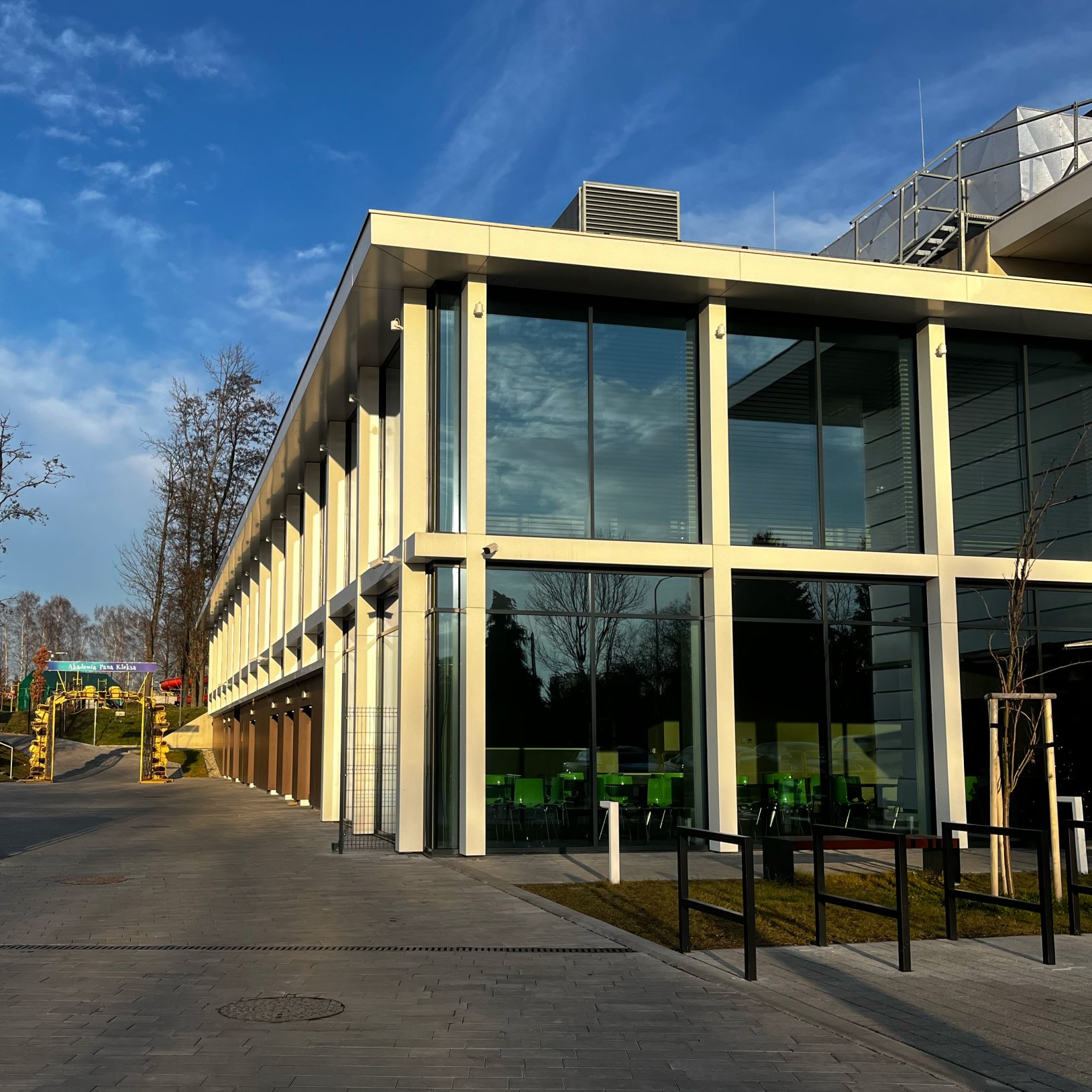 "Kątowy widok nowoczesnego budynku z dużymi szklanymi powierzchniami i białą konstrukcją. W środku widoczne są zielone krzesła, sugerujące obecność sali lub stołówki. Po lewej stronie znajduje się ścieżka prowadząca do placu zabaw z kolorową bramą wejścio