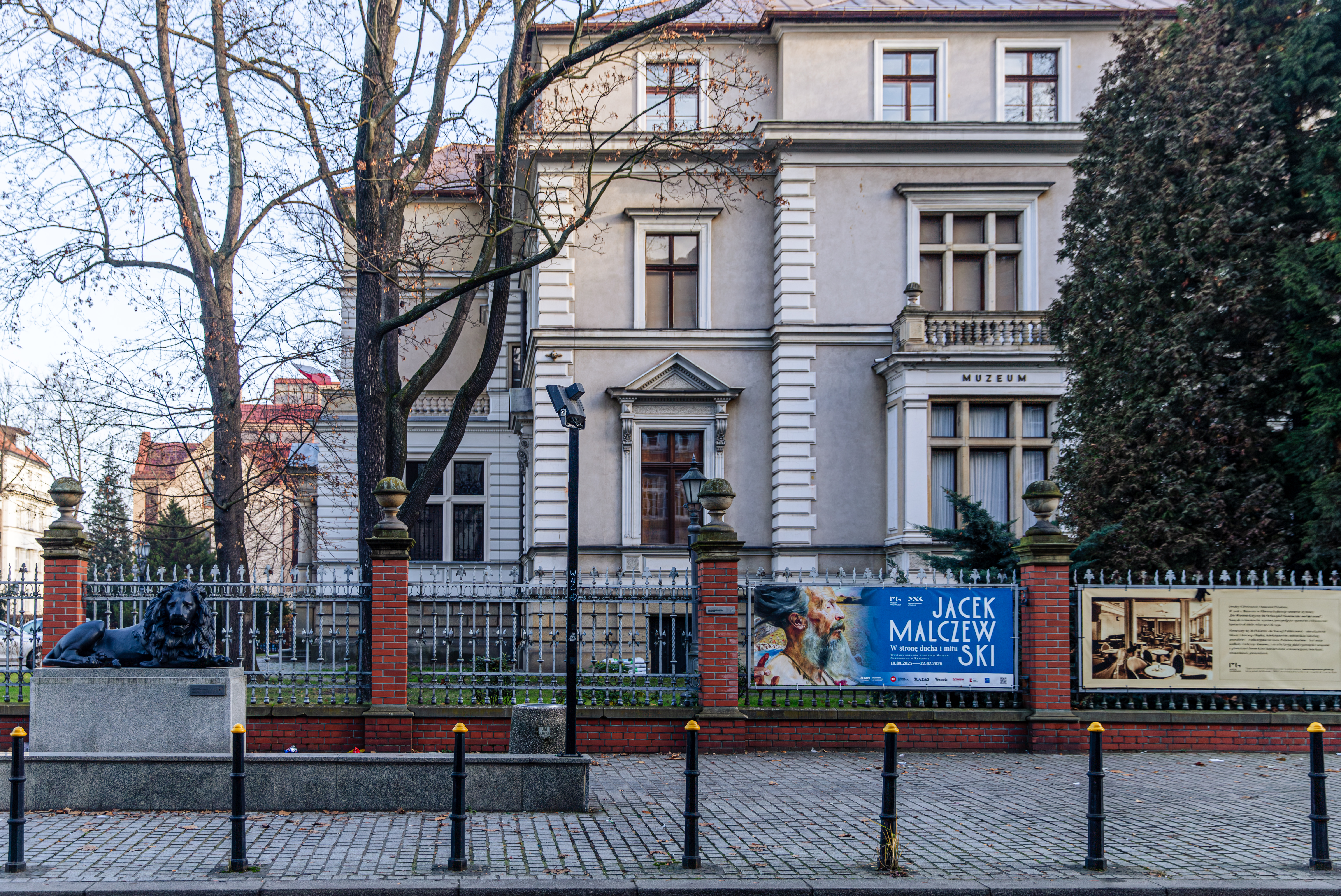 "Widok na front klasycystycznego budynku muzeum, ogrodzonego metalowym płotem z ceglanymi słupkami. Na pierwszym planie pomnik lwa na postumencie oraz baner promujący wystawę Jacka Malczewskiego."