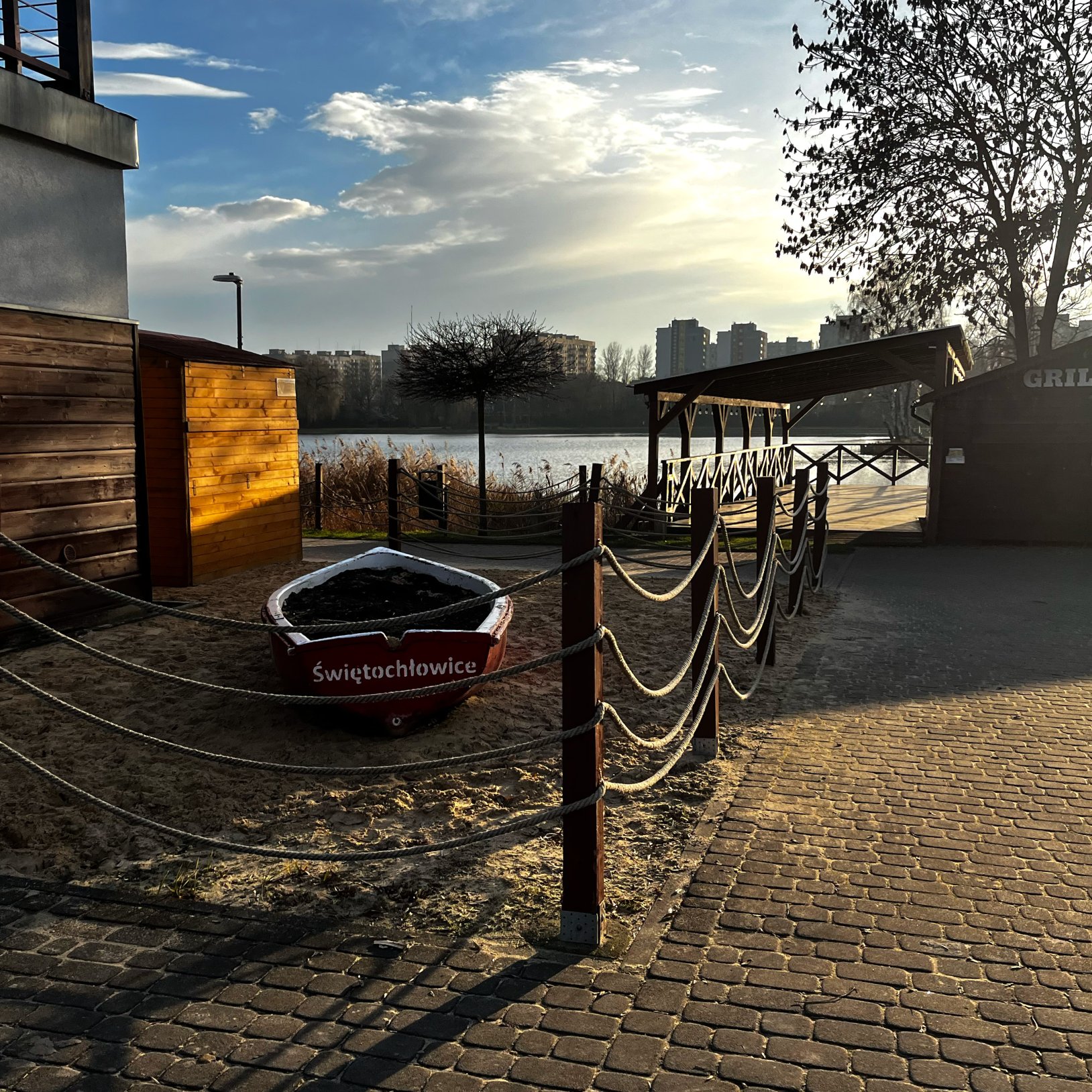 "Widok na drewniany pomost nad jeziorem, częściowo osłonięty zadaszoną wiatą. Obok znajduje się czerwona łódź z napisem 'Świętochłowice', ustawiona na piasku. W tle jezioro, zabudowania mieszkalne i drzewa bez liści. Scena sfotografowana przy zachodzącym