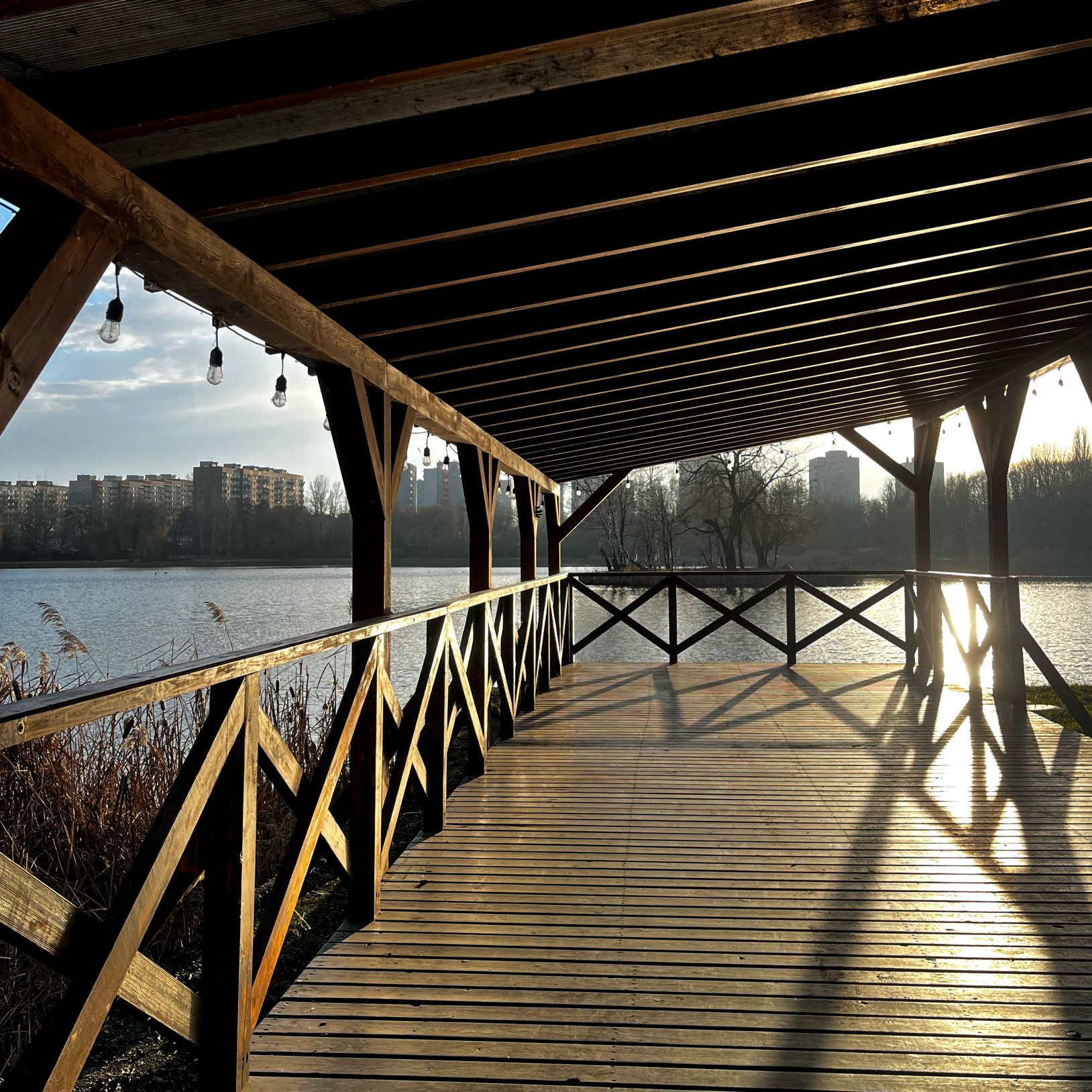 "Zadaszony, drewniany pomost nad jeziorem z balustradami po bokach i wiszącymi żarówkami. Po lewej stronie widoczna tafla wody, w tle zabudowa mieszkaniowa oraz drzewa. Słońce rzuca długie cienie na podłogę konstrukcji."
