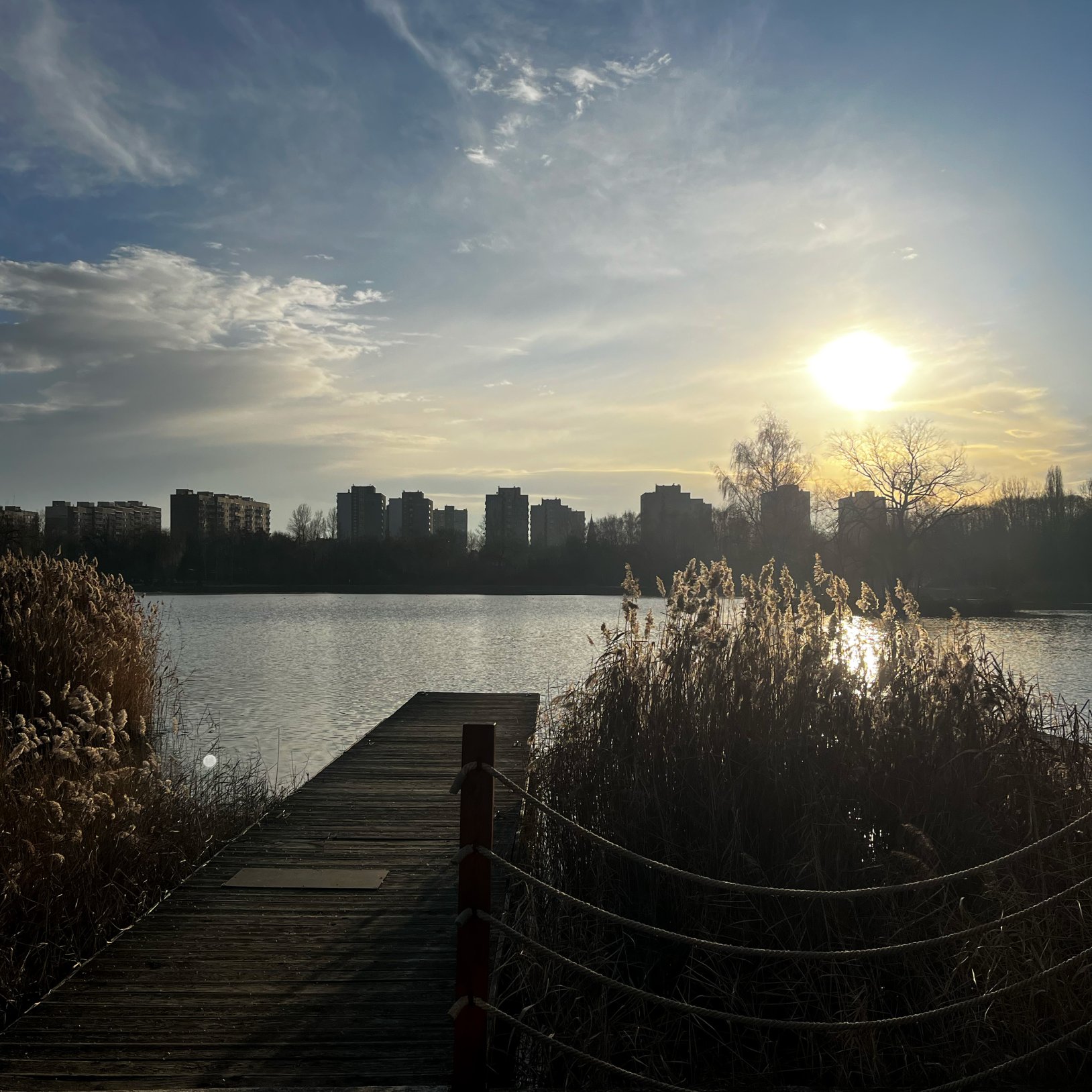 "Widok na drewniany pomost prowadzący na jezioro, otoczony trzcinami. Na horyzoncie widać wysokie bloki mieszkalne na tle zachodzącego słońca. Niebo pokryte częściowo chmurami, a promienie słoneczne odbijają się od tafli wody."