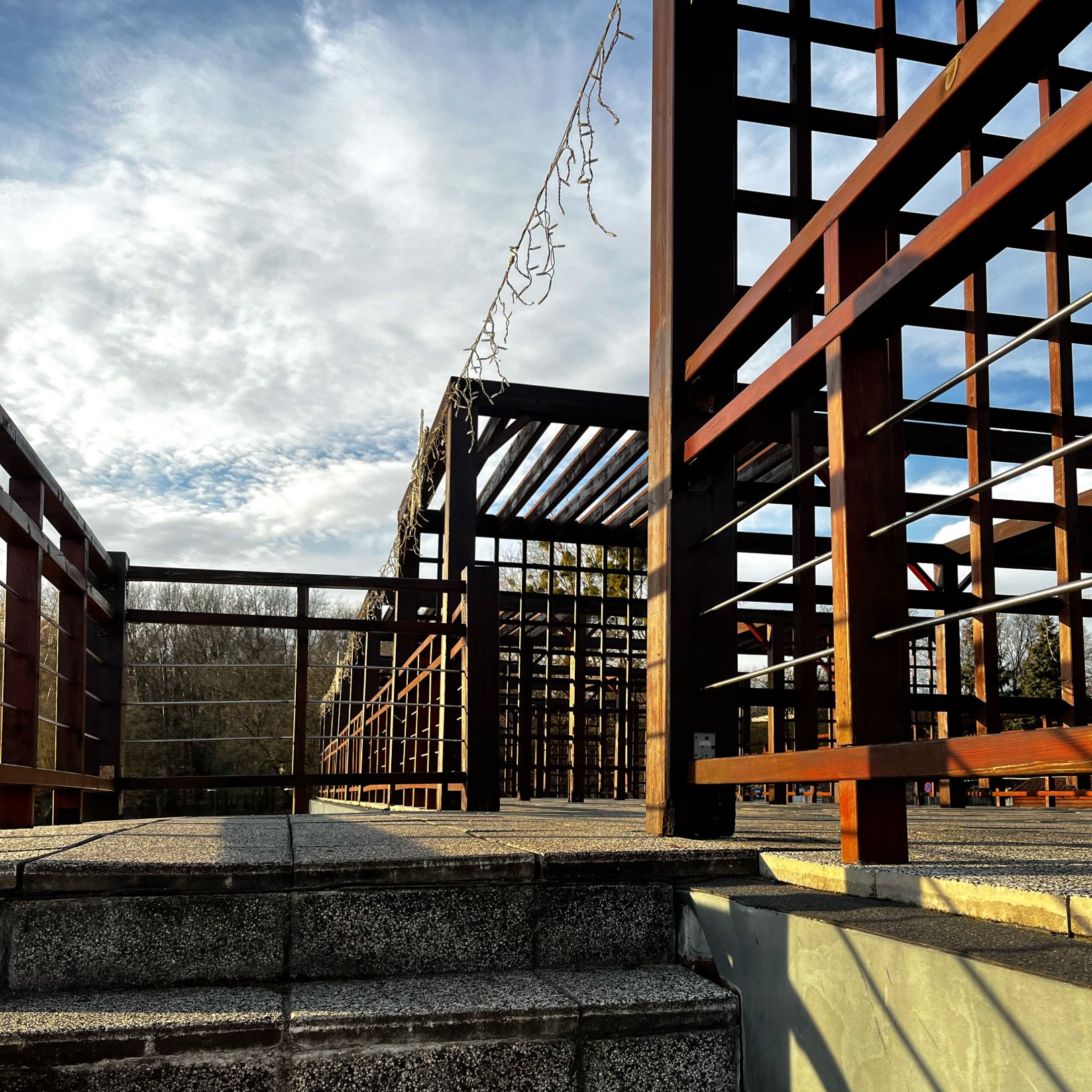 "Drewniana konstrukcja pergoli z kratownicą, ustawiona na betonowej platformie z kilkoma stopniami. Nad konstrukcją rozwieszone są dekoracyjne lampki. W tle widoczne drzewa i niebo z rozproszonymi chmurami."