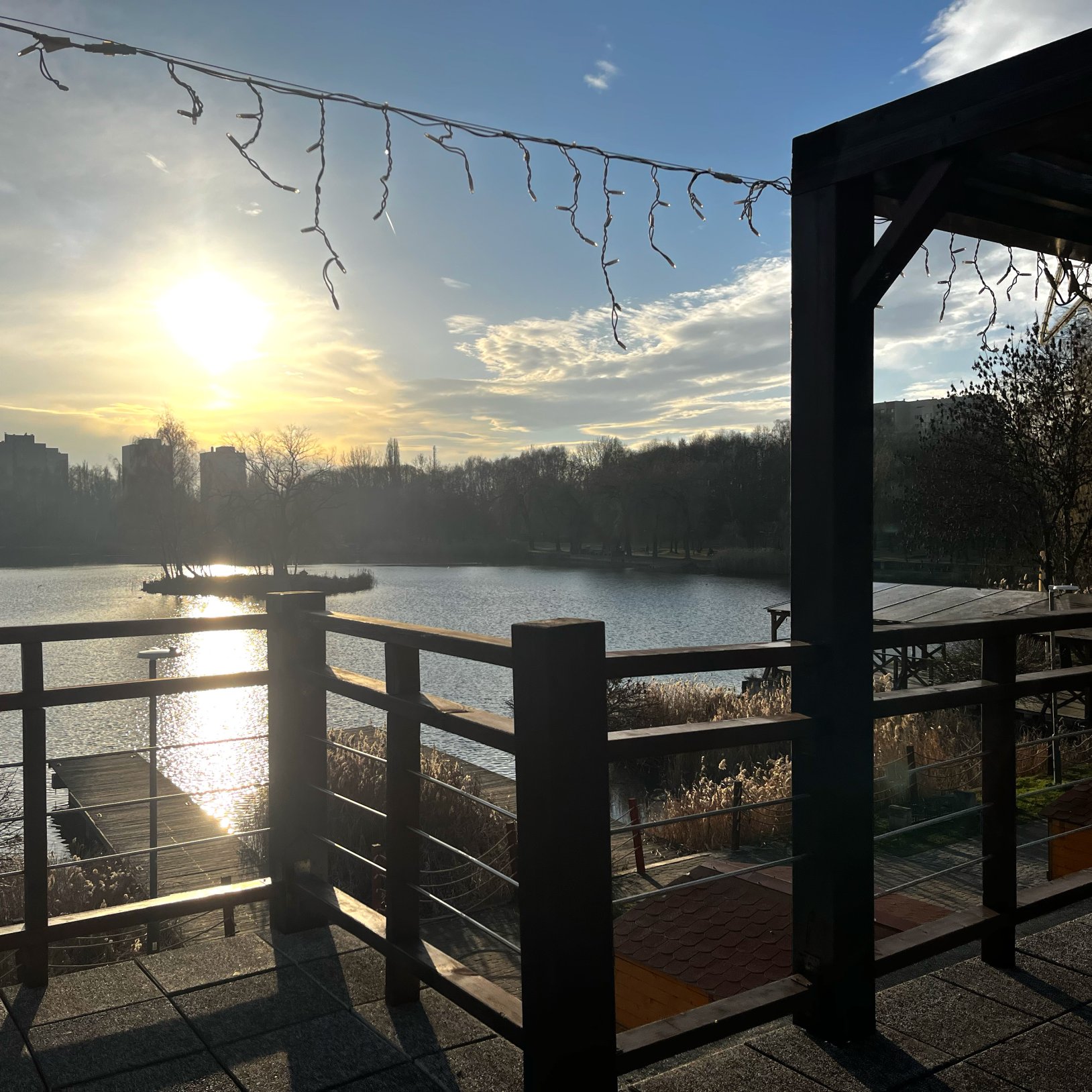 "Widok z podestu z balustradami na jezioro w świetle zachodzącego słońca. Nad głową zawieszone są dekoracyjne lampki. W tle widać linię drzew, pomost oraz wyspę z drzewami. Po lewej stronie widoczna tafla jeziora i trzciny."   }