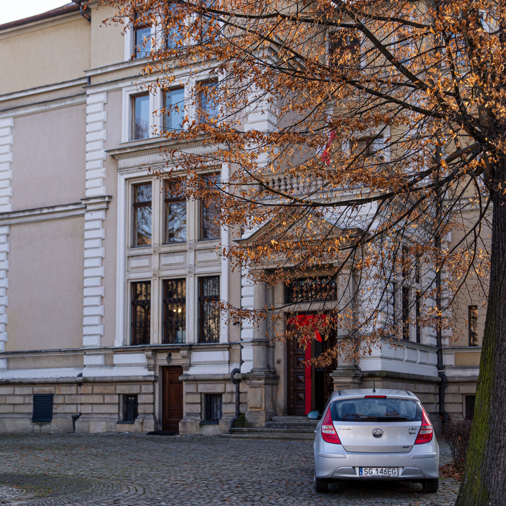 "Zabytkowy budynek z jasną elewacją i białymi zdobieniami, częściowo zasłonięty przez drzewo z suchymi liśćmi. Na brukowanym dziedzińcu zaparkowany jest srebrny samochód osobowy."