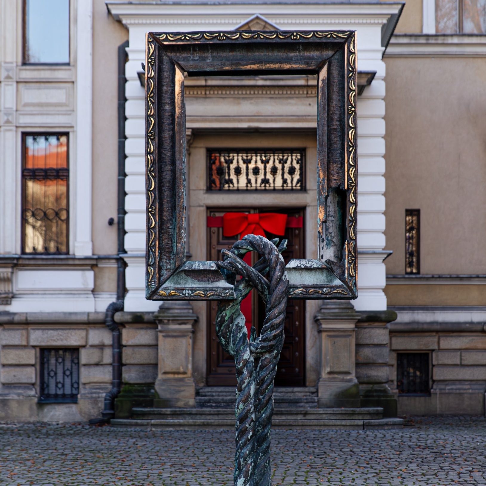 "Rzeźba przedstawiająca skręcony metalowy pręt podtrzymujący dużą ozdobną ramę, ustawiona na brukowanym dziedzińcu przed wejściem do klasycznego budynku. W tle drzwi z czerwoną kokardą i fragmenty elewacji z białymi detalami architektonicznymi."