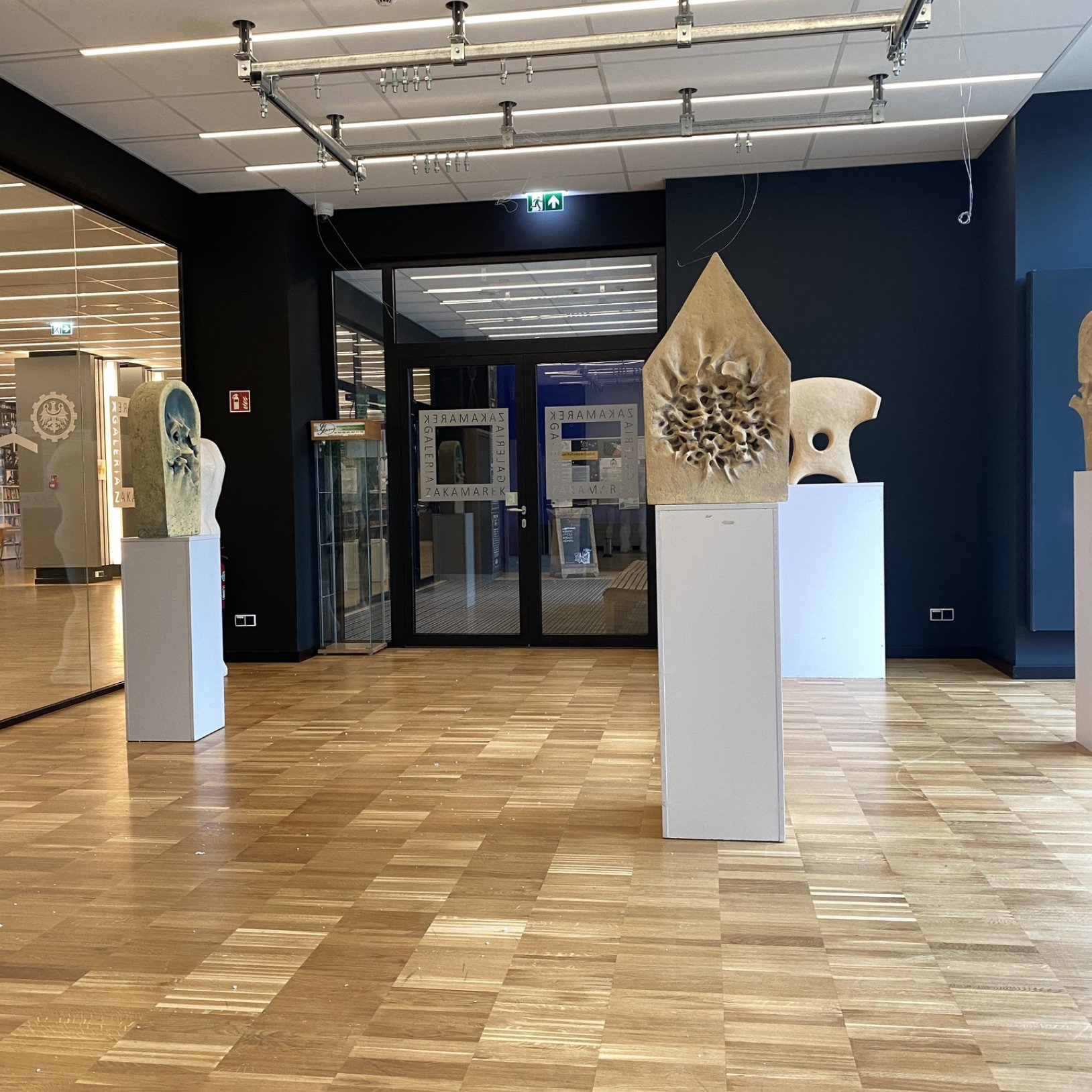 Wnętrze galerii z drewnianą podłogą i ciemnymi ścianami; na białych postumentach stoją jasne rzeźby o abstrakcyjnych kształtach, a w tle widać przeszklone drzwi z napisami ZAKAMAREK oraz oświetlenie sufitowe na metalowych szynach.