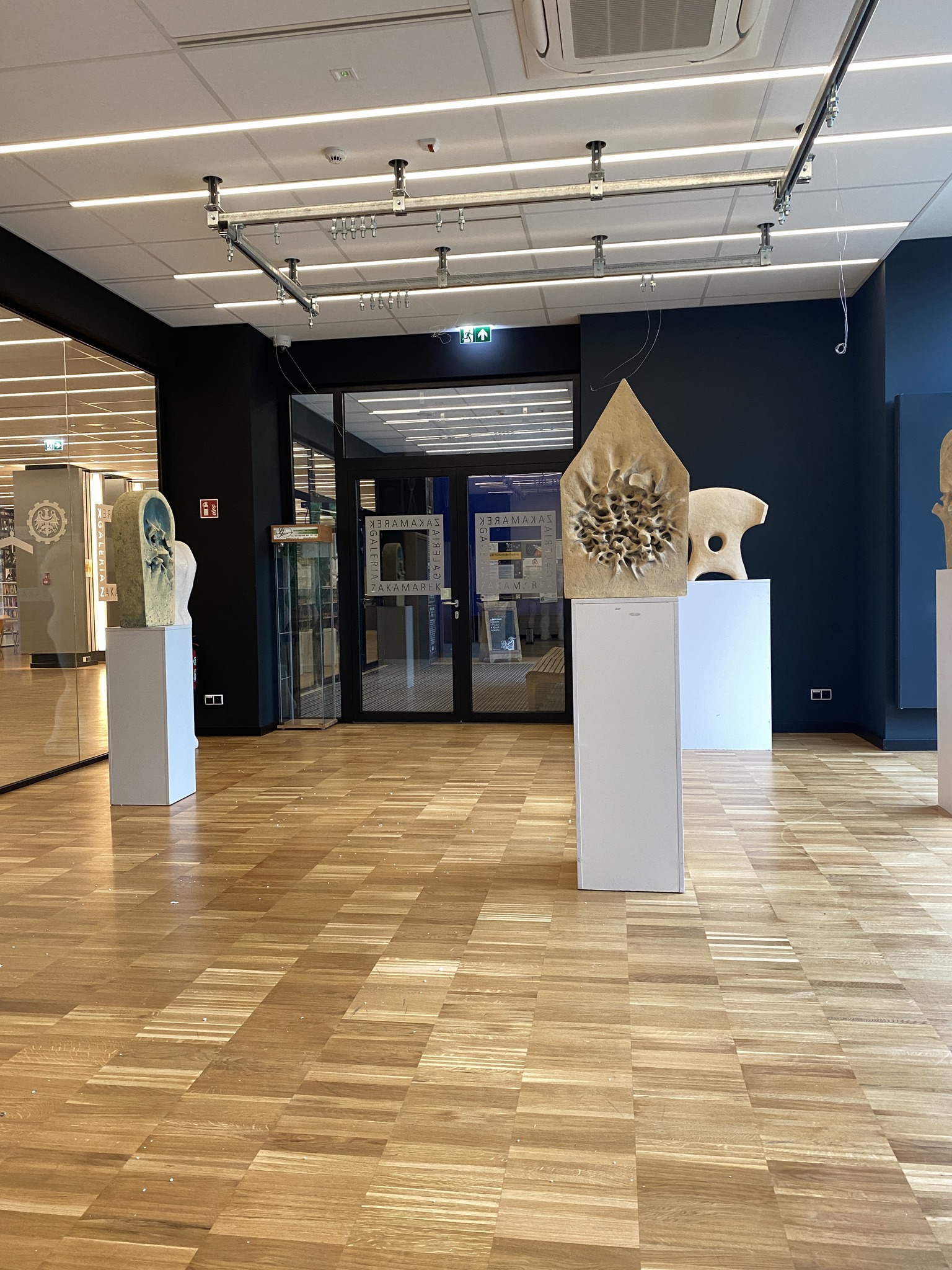Wnętrze galerii z drewnianą podłogą i ciemnymi ścianami; na białych postumentach stoją jasne rzeźby o abstrakcyjnych kształtach, a w tle widać przeszklone drzwi z napisami „ZAKAMAREK” oraz oświetlenie sufitowe na metalowych szynach.