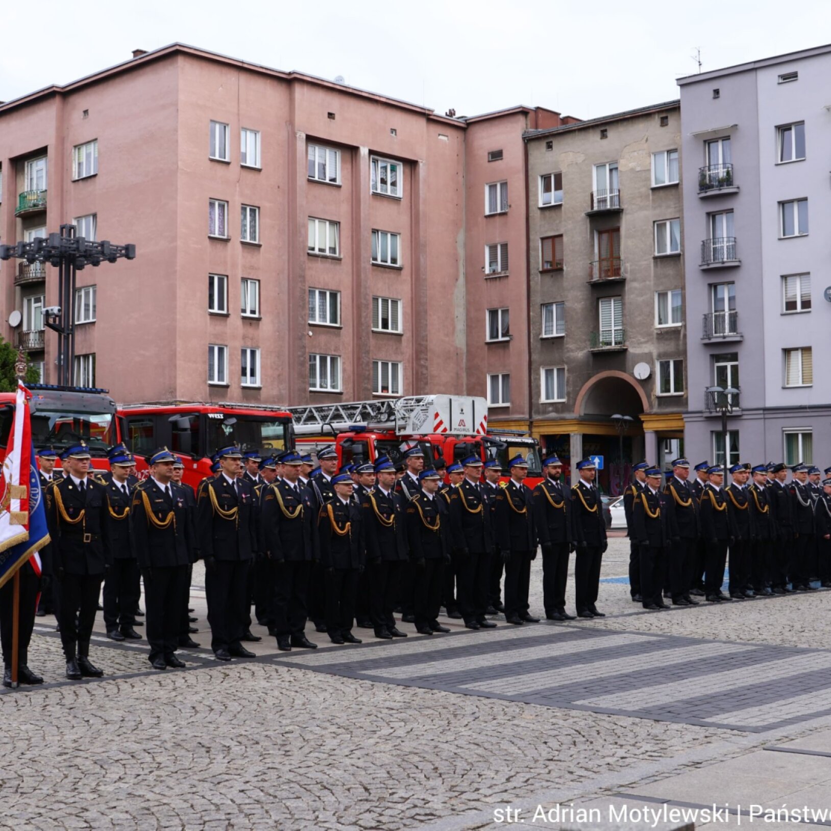 Uroczysty apel strażaków w galowych mundurach ustawionych w szeregu na brukowanym placu przed budynkami, obok wozów strażackich.