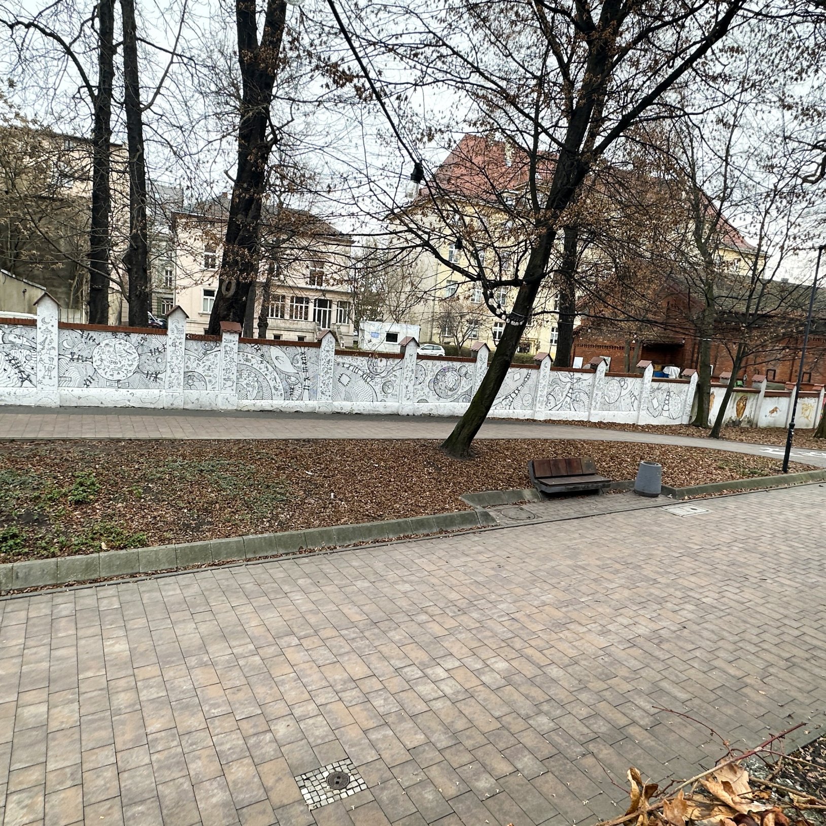 Brukowana alejka w parku z bezlistnymi drzewami; po lewej ciągnie się niski mur z białymi panelami pokrytymi czarno-białymi rysunkami, a w tle widać zabudowania i ogrodzenie.