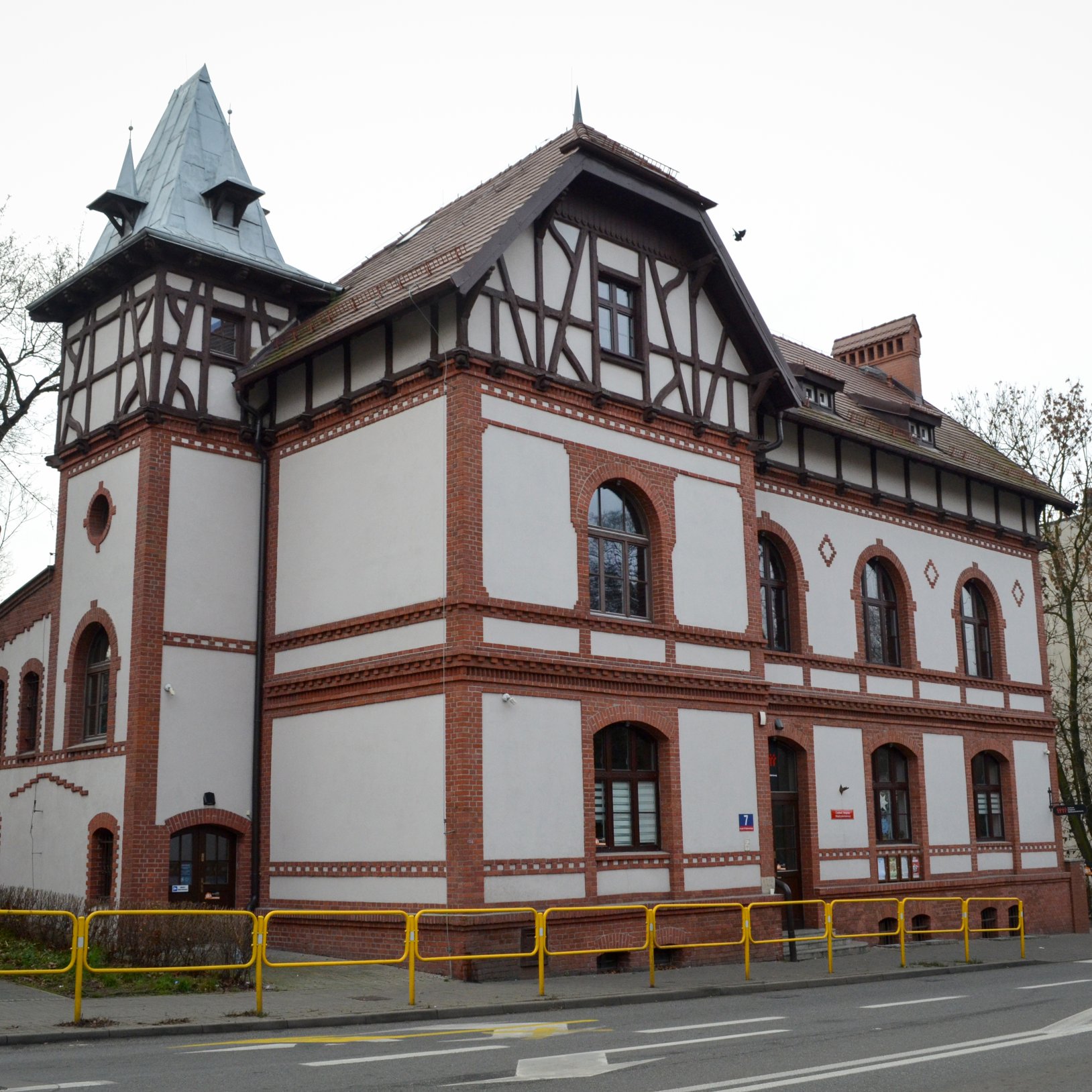 "Zdjęcie przedstawia zabytkowy budynek o jasnej elewacji z czerwonej cegły i tynkiem w stylu muru pruskiego. Budynek ma kilka kondygnacji, duże łukowe okna i wieżyczkę z ostrym, metalowym dachem. Przed budynkiem znajduje się chodnik oddzielony od ulicy żó