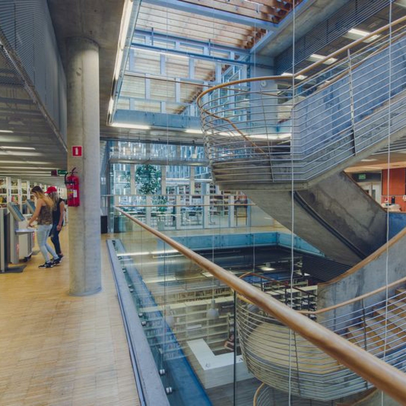 Wnętrze nowoczesnej biblioteki z centralnie umieszczonymi spiralnymi schodami, przeszklonymi barierkami i widocznymi regałami z książkami. Po lewej stronie dwie osoby korzystają z terminali samoobsługowych.