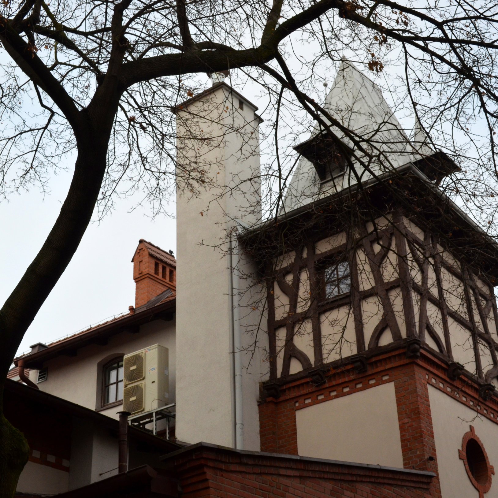 "Fragment elewacji zabytkowego budynku z murem pruskim i charakterystyczną wieżyczką z ostrym, metalowym dachem. W tle widoczny jest komin i gałęzie drzewa bez liści. Na ścianie zamontowana jest jednostka klimatyzacyjna."