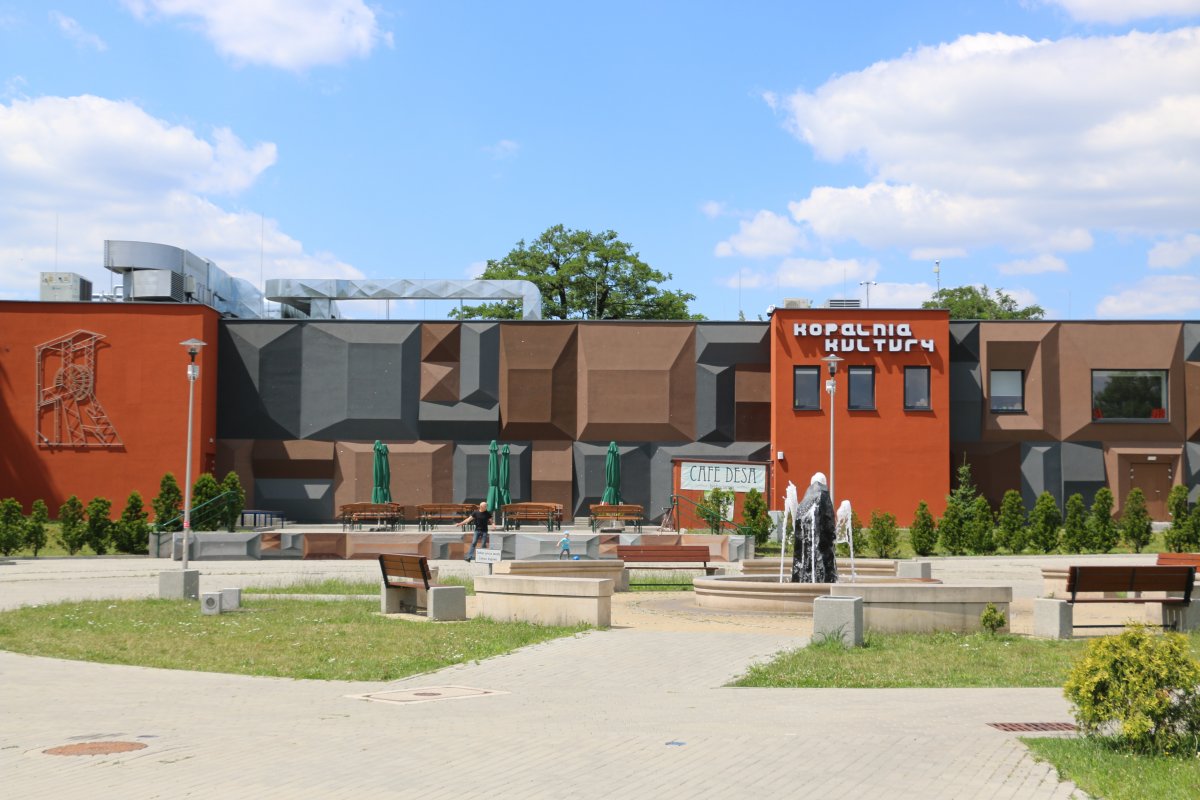 "Widok na nowoczesny budynek Kopalni Kultury w Czeladzi. Elewacja w odcieniach brązu i szarości ma geometryczne, trójwymiarowe formy. Przed budynkiem znajduje się niewielki plac z fontanną, ławkami i ogródkiem kawiarnianym z zielonymi parasolami. Na ścian