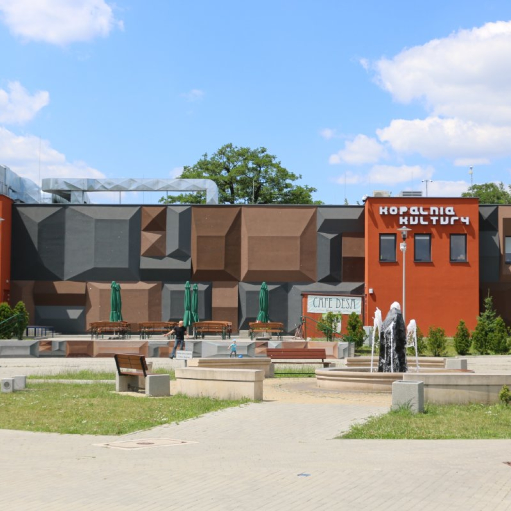 "Widok na nowoczesny budynek Kopalni Kultury w Czeladzi. Elewacja w odcieniach brązu i szarości ma geometryczne, trójwymiarowe formy. Przed budynkiem znajduje się niewielki plac z fontanną, ławkami i ogródkiem kawiarnianym z zielonymi parasolami. Na ścian