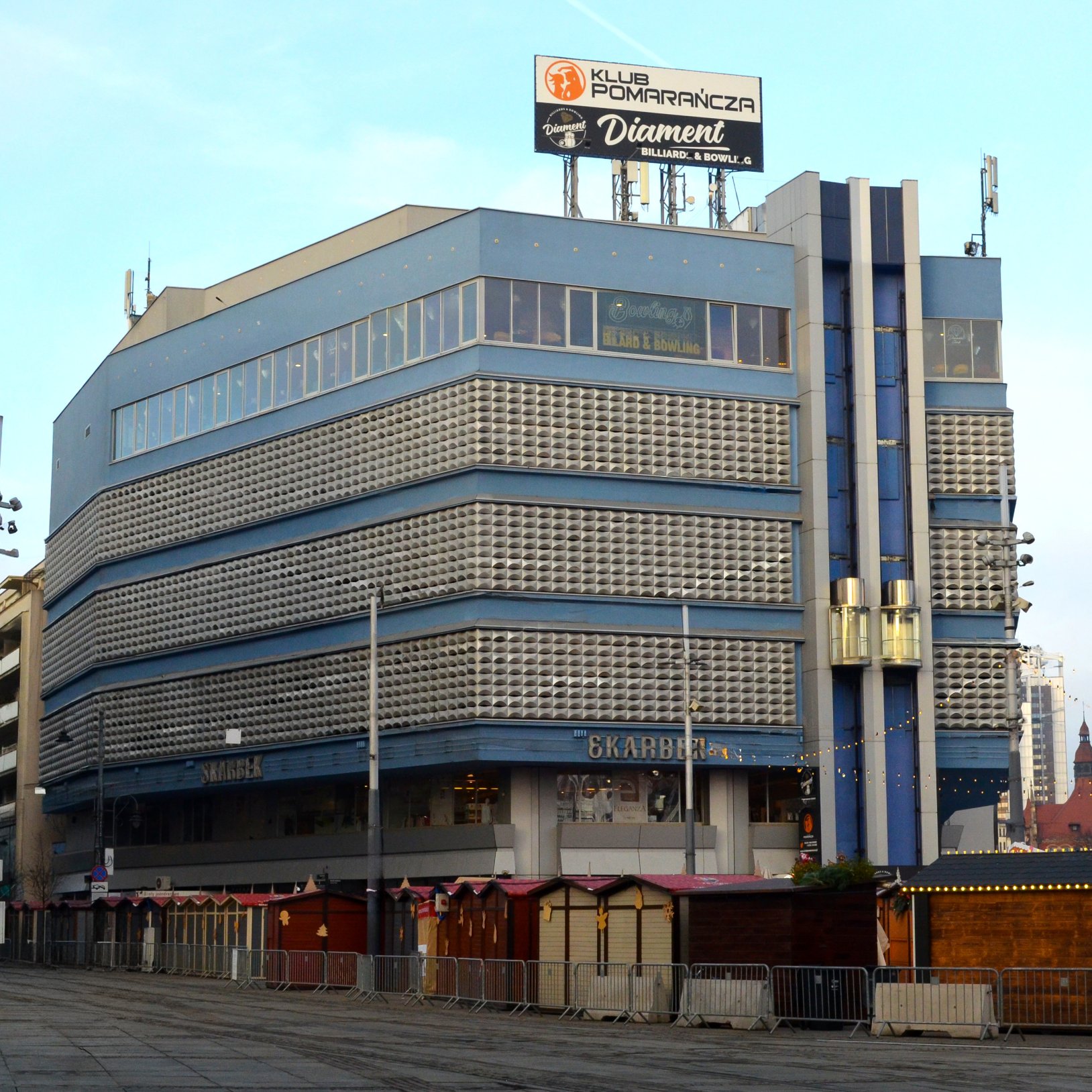 "Zdjęcie budynku handlowego o nazwie 'Skarbek' w Katowicach. Bryła ma charakterystyczną fasadę z elementami kratownicy i niebieskimi akcentami. Na dachu widoczne są szyldy reklamujące Klub Pomarańcza i Diament Billiard & Bowling.