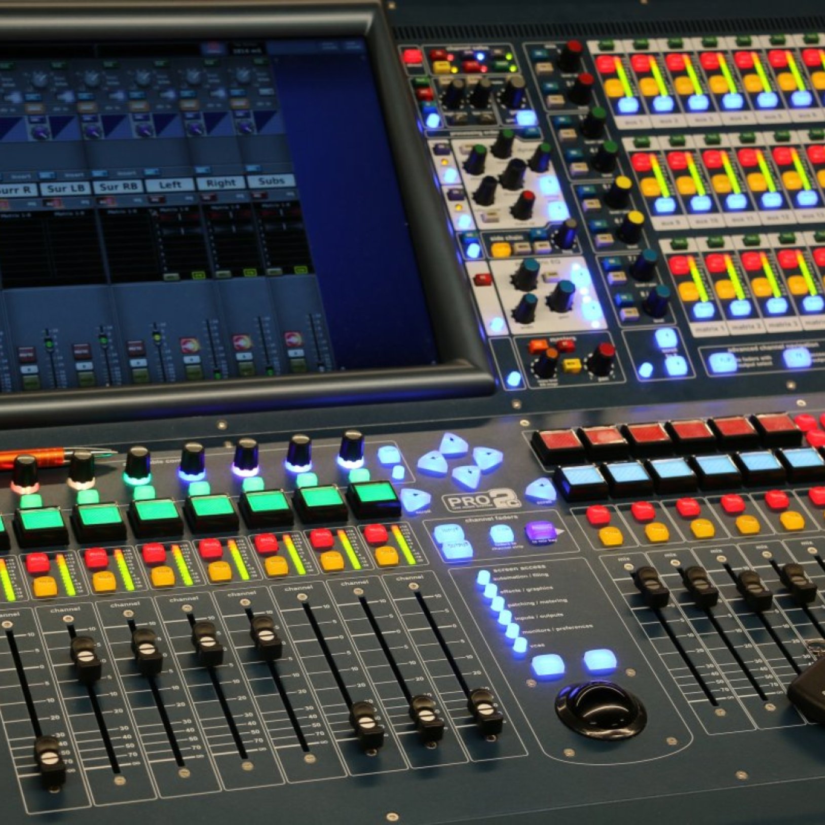 "Zbliżenie na profesjonalną cyfrową konsoletę dźwięku z licznymi suwakami, przyciskami, pokrętłami oraz kolorowymi podświetleniami. W centralnej części znajduje się ekran dotykowy z wizualizacją kanałów audio. Sprzęt oznaczony jest logo PRO2."