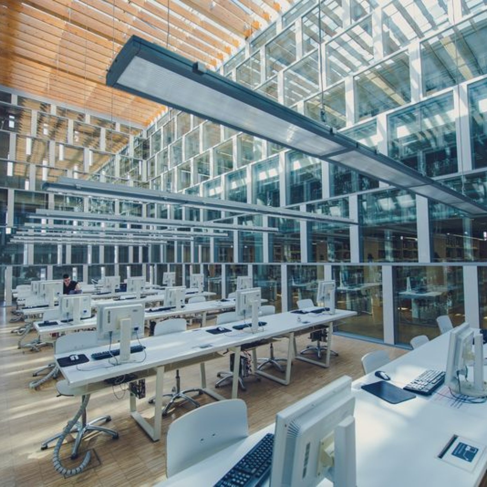 Nowoczesna przestrzeń biblioteczna z licznymi komputerami na białych biurkach. Wnętrze jest dobrze oświetlone dzięki przeszklonym ścianom i sufitowi z drewnianymi belkami.