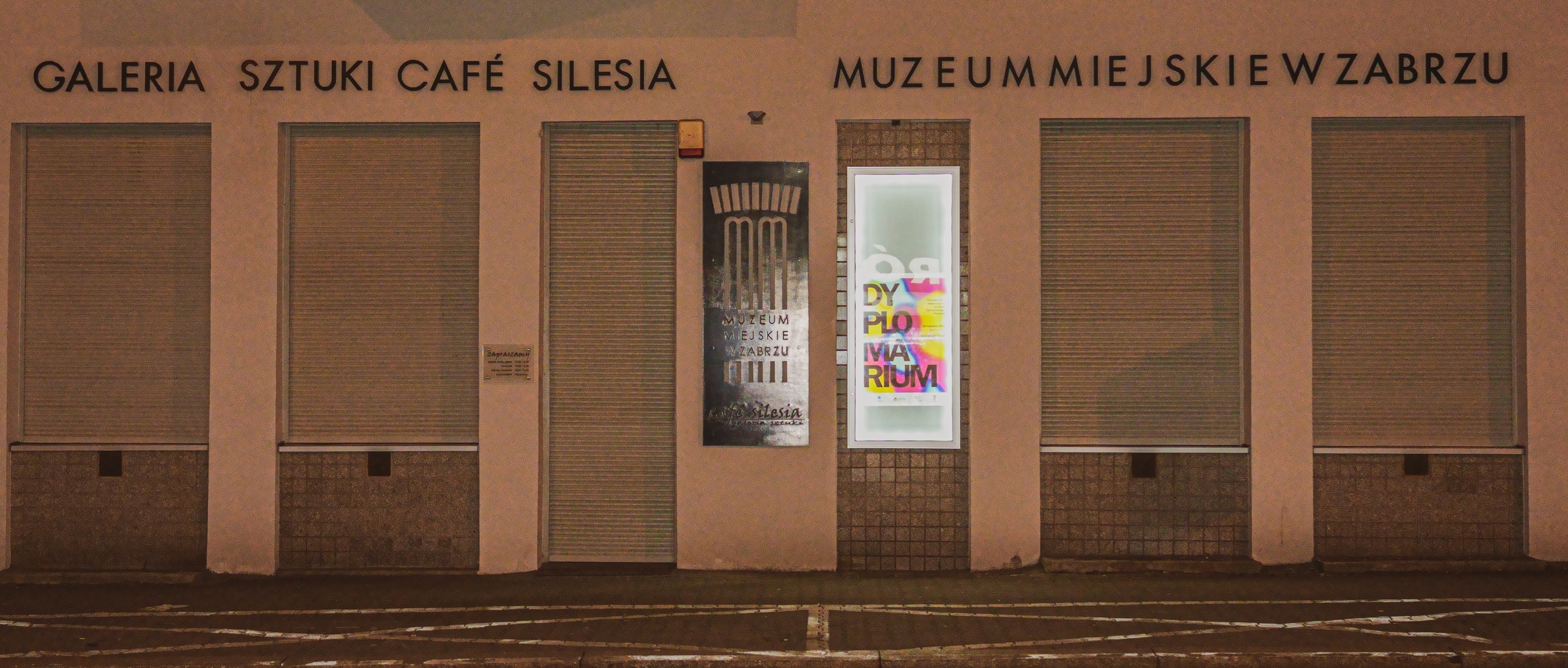 "Nocne zdjęcie fragmentu elewacji budynku Muzeum Miejskiego w Zabrzu – Galerii Sztuki Café Silesia. Na ścianie widoczna jest podświetlona gablota z kolorowym plakatem oraz metalowa tablica z logo muzeum. Otoczenie jest ciemne, a szyby zabezpieczone roleta