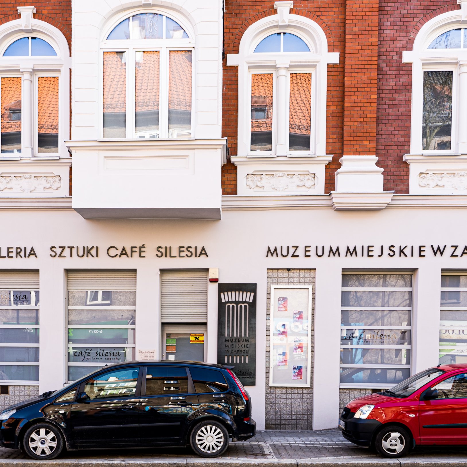 "Dzienny widok na elewację Muzeum Miejskiego w Zabrzu – Galerię Sztuki Café Silesia. Budynek ma biało-czerwoną fasadę z ozdobnymi gzymsami i dużymi, łukowatymi oknami na piętrze. Przed wejściem zaparkowane są dwa samochody osobowe."