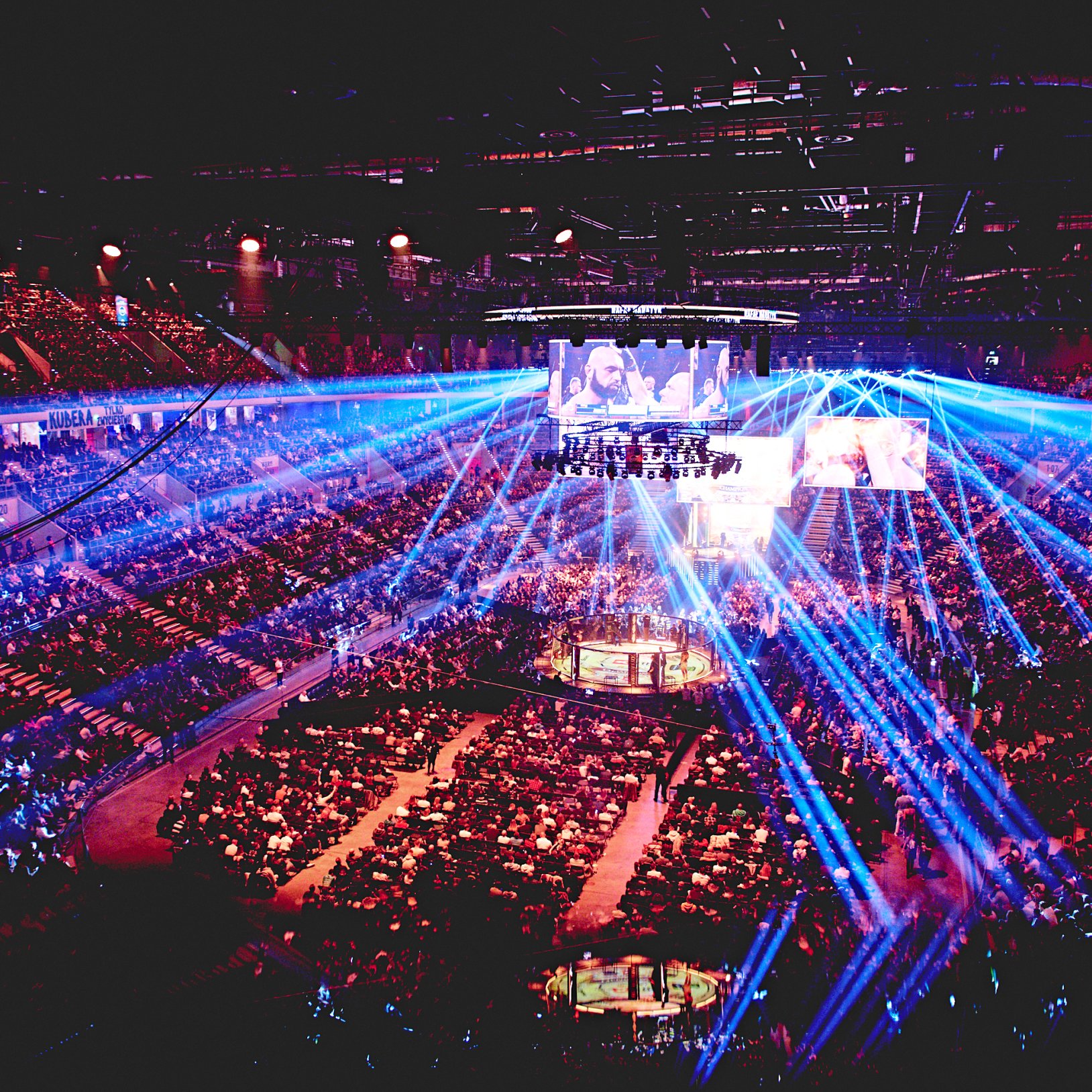 "Widok wnętrza PreZero Areny Gliwice podczas dużego wydarzenia, prawdopodobnie gali sportów walki. Tłum ludzi wypełnia trybuny i przestrzeń wokół oktagonu. Z sufitu emitowane są liczne promienie świetlne w odcieniach niebieskiego i fioletu, tworząc efekto