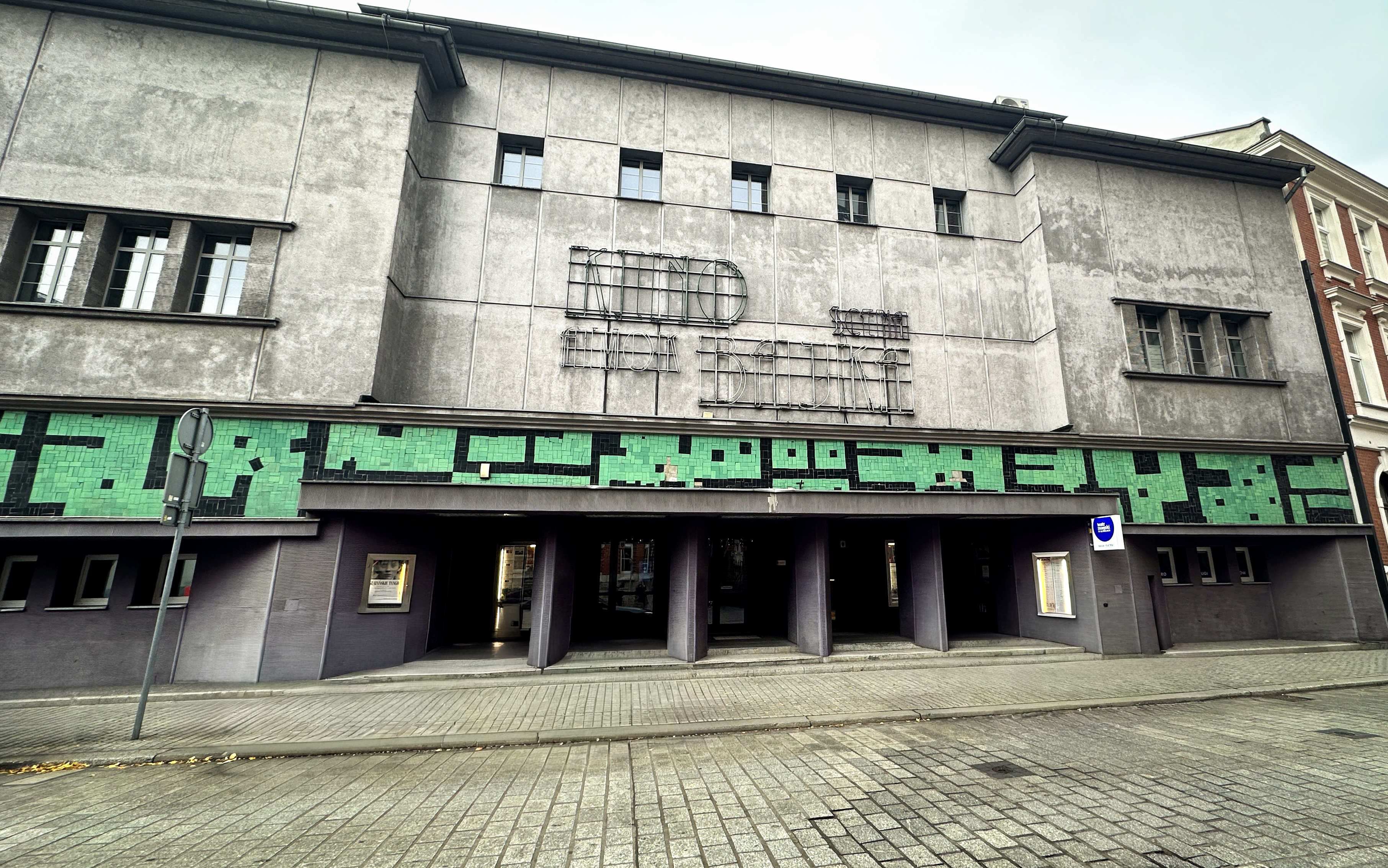 "Fasada budynku kina z dużym napisem neonowym 'KINO AMOK' na szarej, betonowej elewacji. Nad wejściem widoczny zielony pas dekoracyjny z mozaikowych płytek. Wejście do kina składa się z kilku ciemnych, otwartych przejść. Przed budynkiem brukowany chodnik