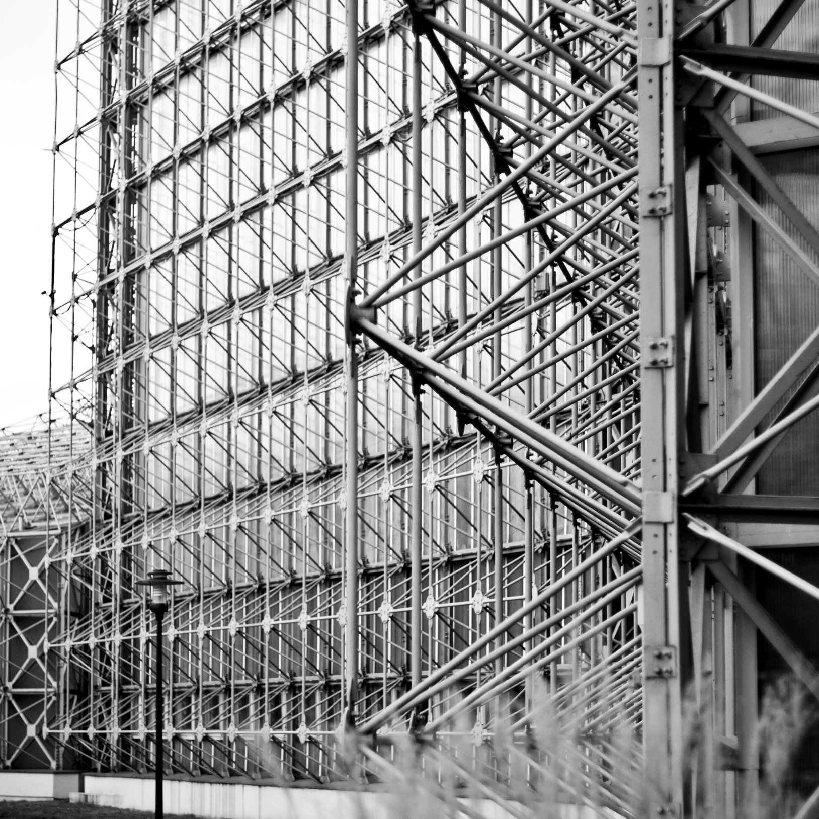 "Czarno-białe zdjęcie fragmentu nowoczesnego budynku z fasadą z metalowej siatki konstrukcyjnej i przeszkleń. Konstrukcja składa się z gęsto rozmieszczonych rur i wsporników, tworząc geometryczny wzór."