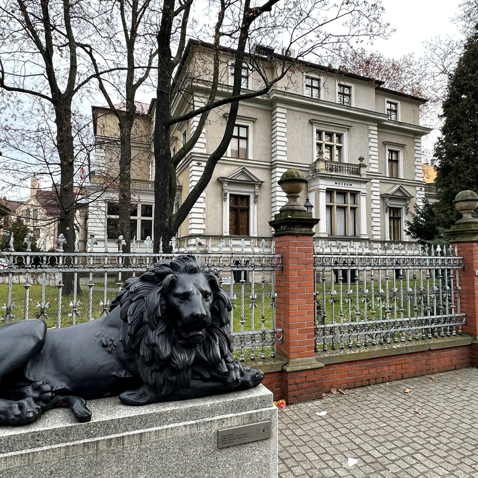 "Widok na zabytkową Willę Caro w Gliwicach, sfotografowaną od frontu. Budynek ma jasną elewację, dekoracyjne detale architektoniczne i drewniane drzwi wejściowe z kolumnowym portalem. Przed budynkiem widoczne są zaparkowane samochody, roślinność i nowocze