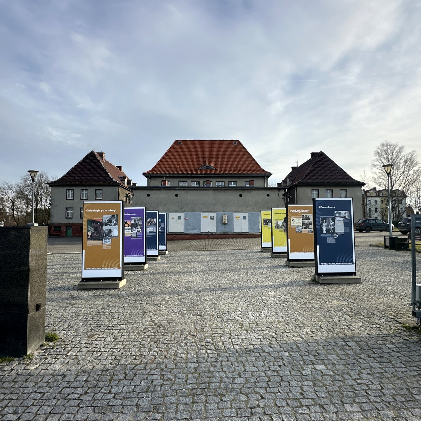 "Plac z wystawą plenerową składającą się z plansz informacyjnych ustawionych w dwóch rzędach na brukowanej nawierzchni. W tle budynek z czerwonym dachem i szarymi ścianami, po bokach dwa mniejsze budynki o podobnym stylu. Wystawa odbywa się na otwartej pr