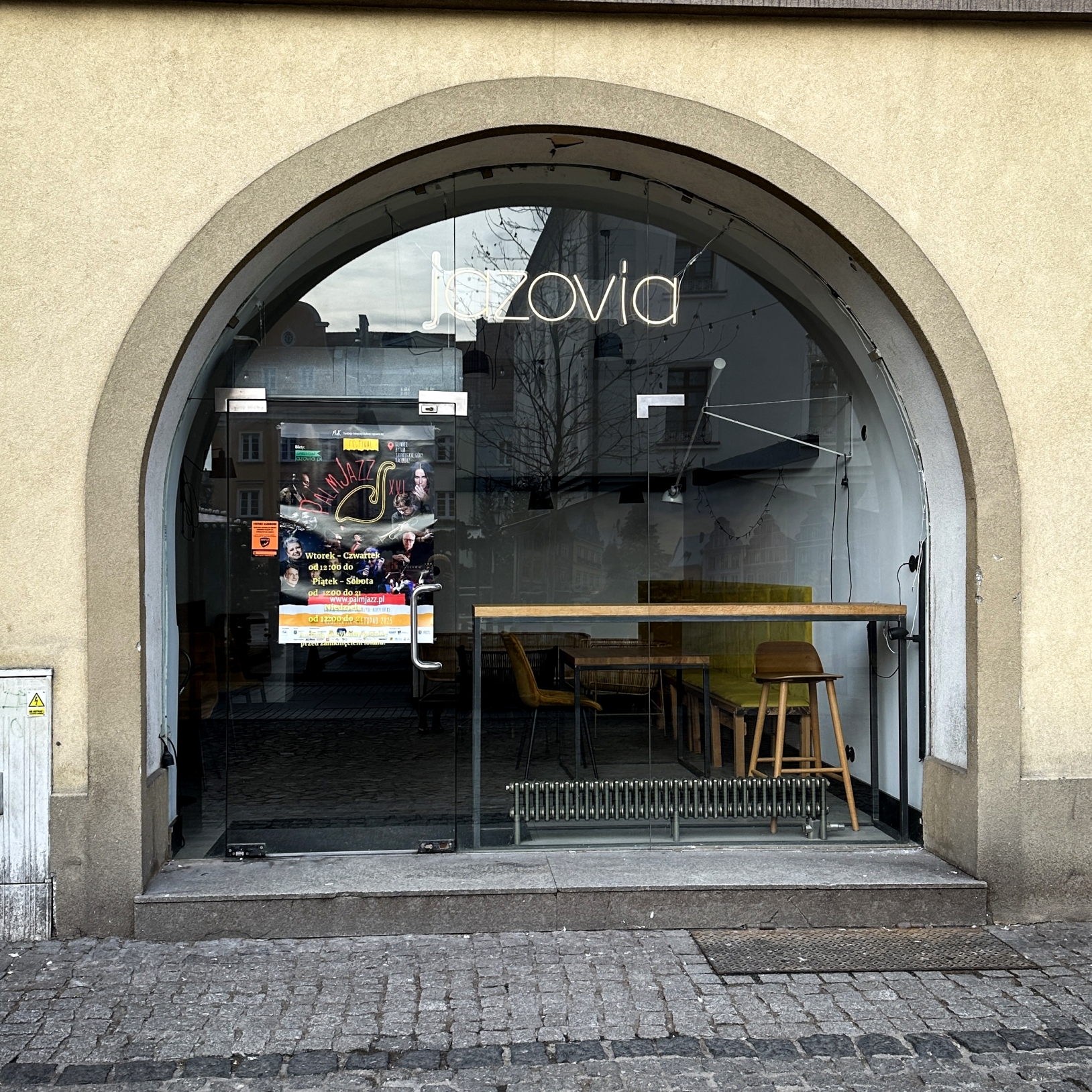 "Wejście do lokalu o nazwie 'Jazovia' z dużą, przeszkloną witryną w kształcie łuku. Na drzwiach znajduje się kolorowy plakat promujący wydarzenie muzyczne. Wnętrze widoczne przez szybę zawiera drewniany bar, krzesła i stoliki. Otoczenie sugeruje lokal zna