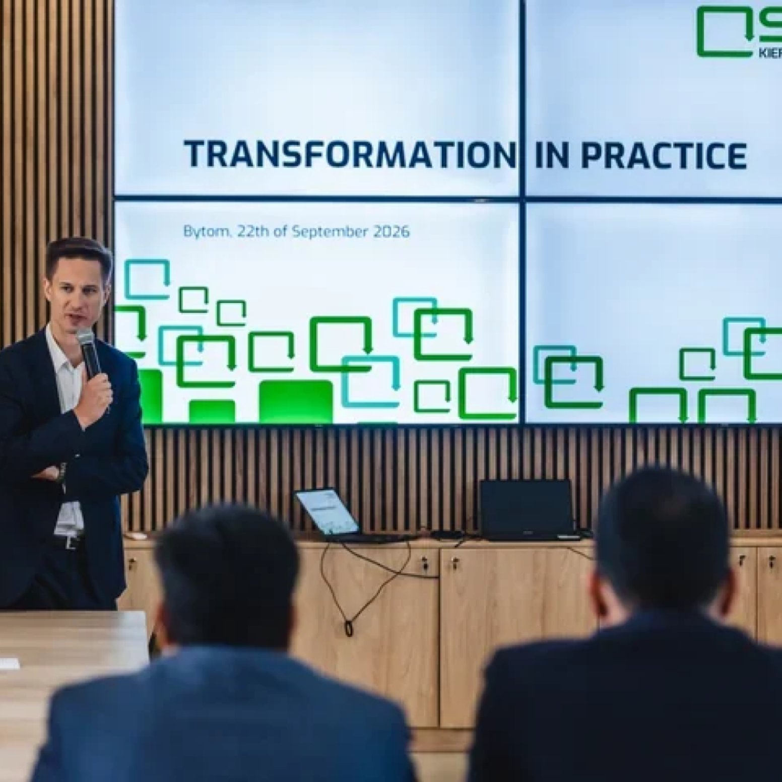 Mówca z mikrofonem przed ekranem z białym tłem i zielonym napisem Transformation in practice