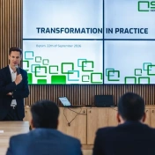 Mówca z mikrofonem przed ekranem z białym tłem i zielonym napisem Transformation in practice