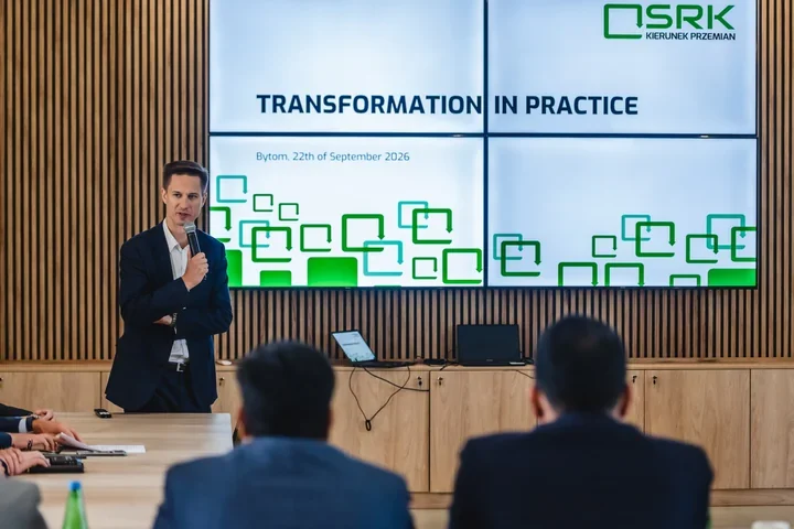 Mówca z mikrofonem przed ekranem z białym tłem i zielonym napisem "Transformation in practice"