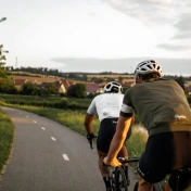 dwóch rowerzystów w kaskach jadących po asfaltowej drodze na tle pola